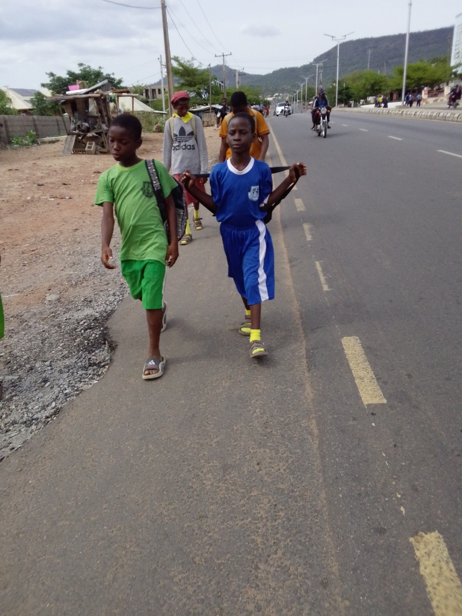 The image shows a group of young boys walking along the side of a road in Adankolo, Nigeria. The boy in front wears a green shirt and shorts with sandals, carrying a backpack. Another boy, dressed in a blue soccer uniform with yellow socks and sandals, is slightly behind him, also carrying a backpack. Further back, other people are visible, including one wearing an Adidas shirt. A road stretches beside them, with vehicles, including a motorcycle, passing by. In the background, there are buildings, trees, and a mountain. A small, dilapidated structure stands on the side of the road, next to a dirt patch. The sky is overcast.