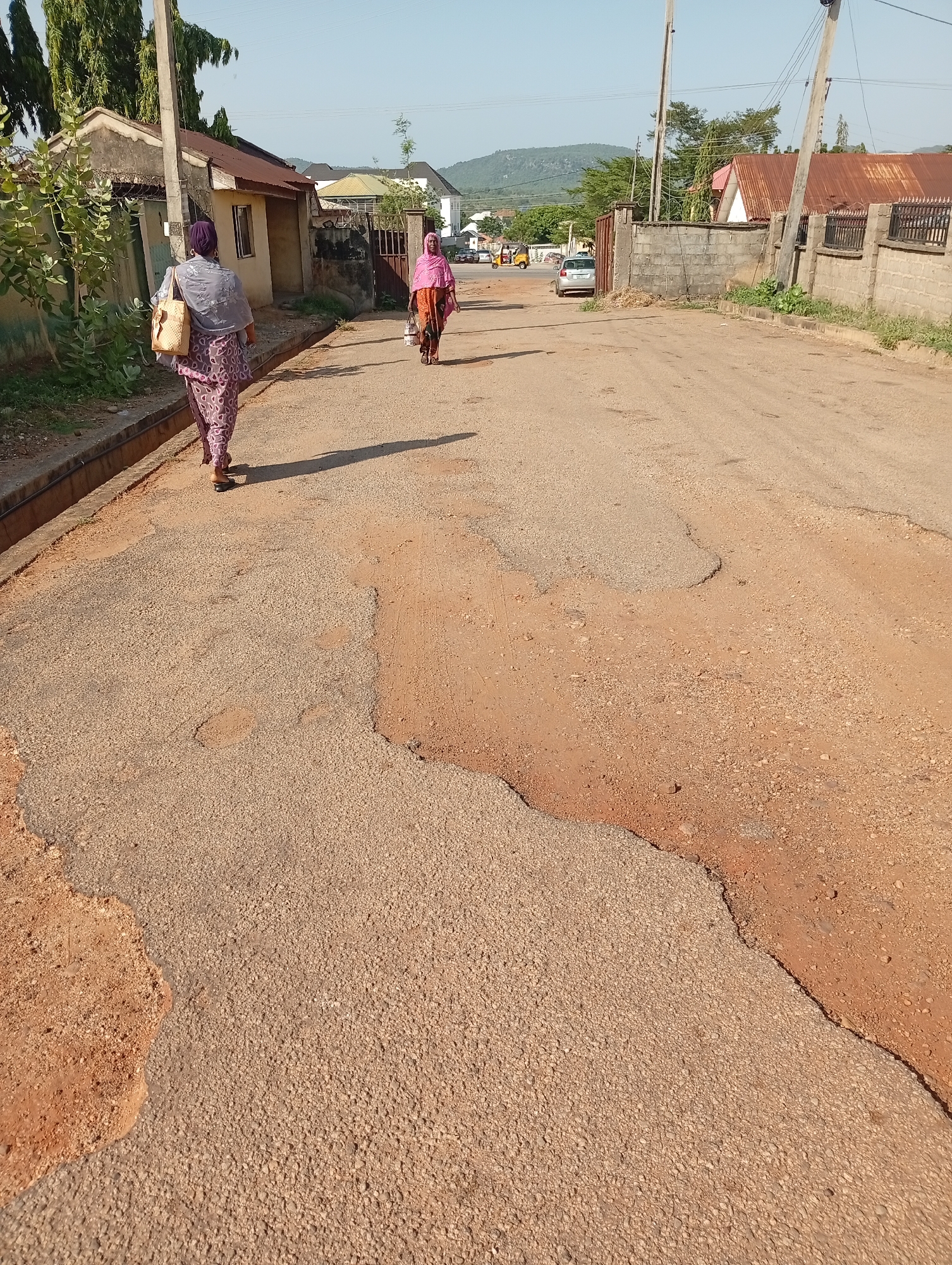 The image shows a deteriorated asphalt road in Adankolo, Nigeria. Two women walk away from the camera along the right side of the road. The woman closer to the camera wears a patterned skirt, a grey head covering, and carries a tan bag over her shoulder. The second woman wears a pink head covering and a patterned dress. The road has extensive patches of missing asphalt exposing underlying dirt. Buildings and fences line both sides of the road. In the background, a silver car is parked on the left side of the road near an intersection. A hill is visible in the distance. The sky is clear and the lighting suggests it is daytime.