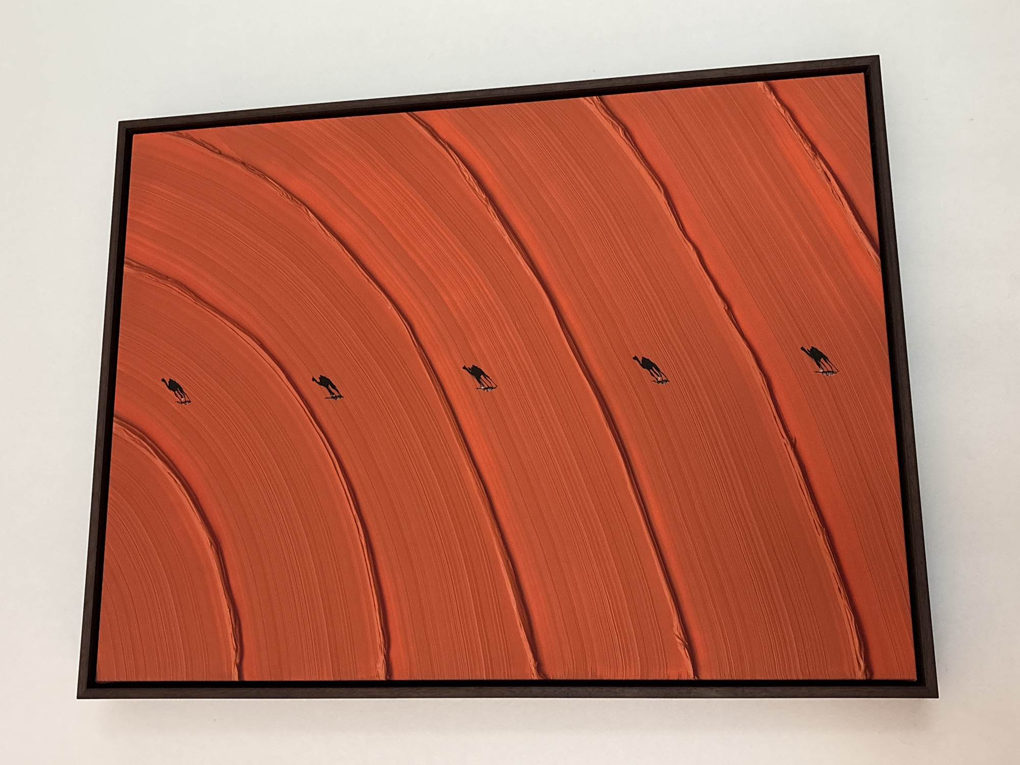 The image displays a framed abstract artwork featuring a vibrant orange background with layered brushstrokes that create a sense of undulating waves or sand dunes. Scattered across these orange layers are small, silhouetted figures that appear to be walking or running, adding a human element to the abstract composition. The artwork is framed in a dark, thin border and is mounted on a white wall, suggesting it is displayed indoors, likely in a gallery, home, or office setting. There is no visible indication of the city of Dubai, United Arab Emirates.
