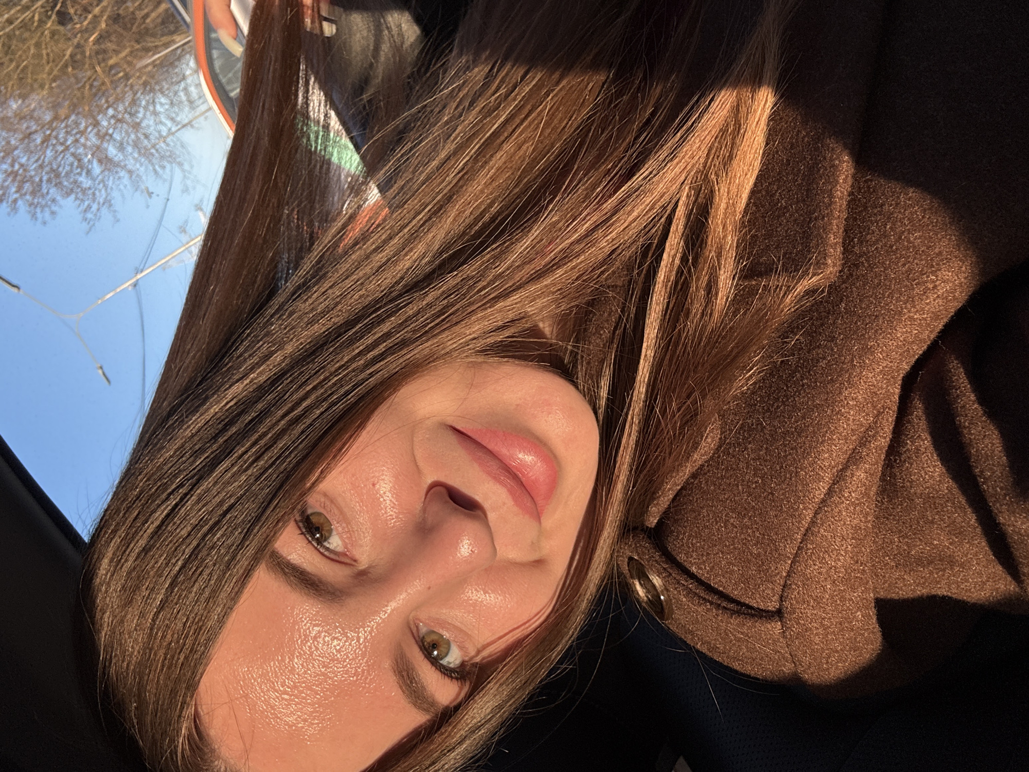 A woman with long, brown hair is pictured from a close-up angle. She has light skin, green eyes, and is wearing a brown coat. Part of a blue sky and some trees are visible in the background. No specific information can be inferred regarding a location in the city of Likino-Dulyovo, Russia.