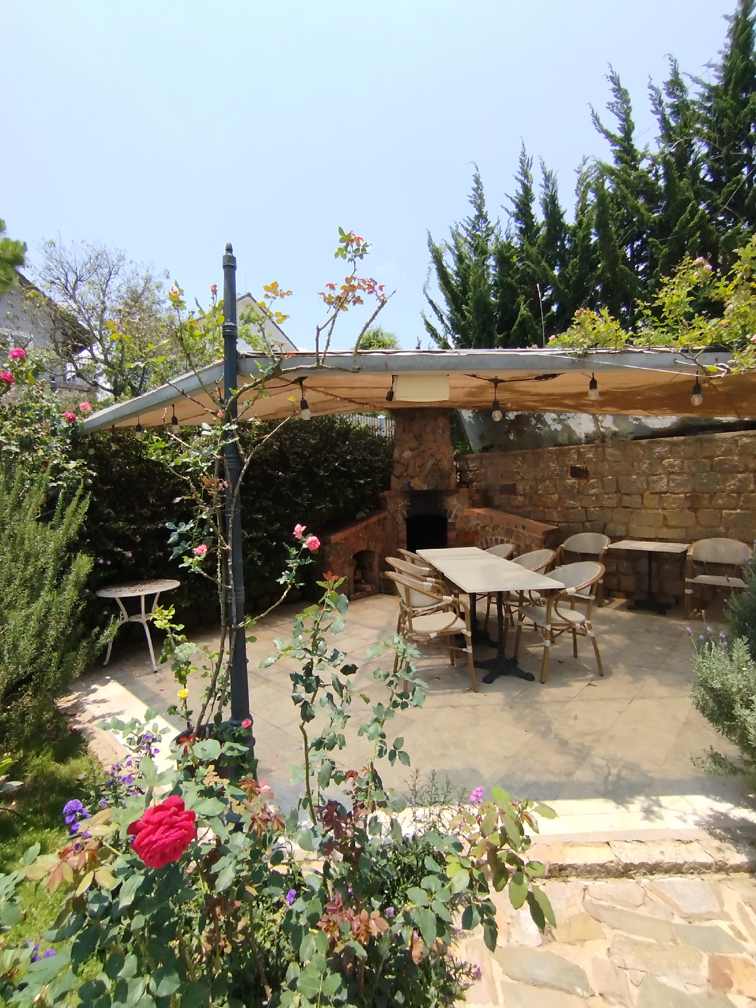 The image shows a courtyard in Lang Biang - Đà Lạt, Vietnam. A stone patio is furnished with a dining table and chairs under a cloth awning, which is supported by a lamp post. A brick and stone fireplace/barbecue area is against the back wall. Vegetation, including roses, rosemary, and other plants, surrounds the patio area. A small, white table sits on the edge of the patio. The scene appears to be outdoors on a sunny day. Tall evergreen trees are visible in the background.