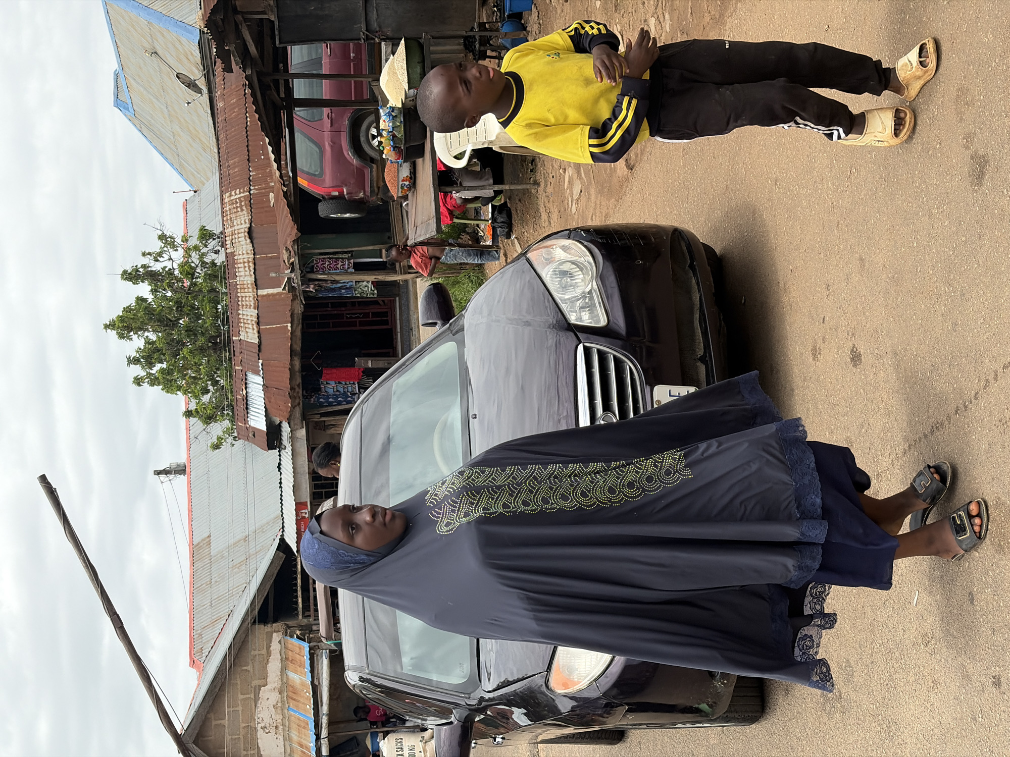 In an outdoor urban setting in Jos, Nigeria, a dark-colored sedan is parked on a dirt road, partially obscuring a young boy wearing a yellow long-sleeved shirt with black and yellow stripes on the sleeves and black pants. He is standing to the right of the car, looking to his left with his mouth slightly open. To the left of the car, a young woman is walking away from the camera, wearing a dark blue hijab that covers her head and extends down to her ankles, adorned with embroidered patterns on the front. She is also wearing dark blue footwear. In the background, several buildings with corrugated metal roofs are visible, with some appearing to be market stalls displaying clothing and goods. Another child in a pink shirt is standing near a table laden with food items, possibly in a market stall. The sky is overcast with clouds.