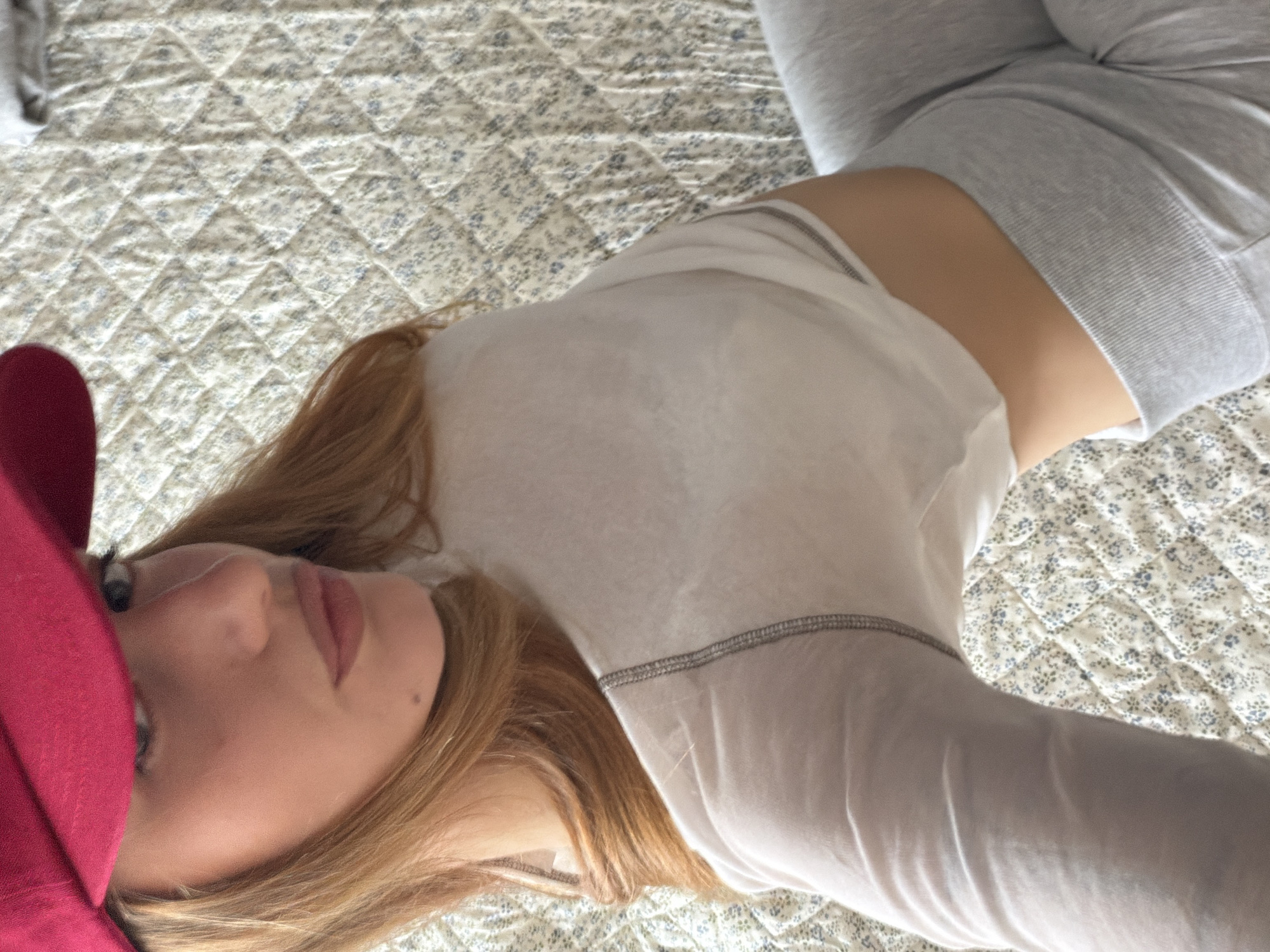 A woman with blonde hair is lying on a bed. She is wearing a red sleep mask and a white long-sleeved shirt. She is also wearing gray sweatpants. The bed has a patterned quilt. There is no evidence to suggest this photo was taken in Sofia, Bulgaria.