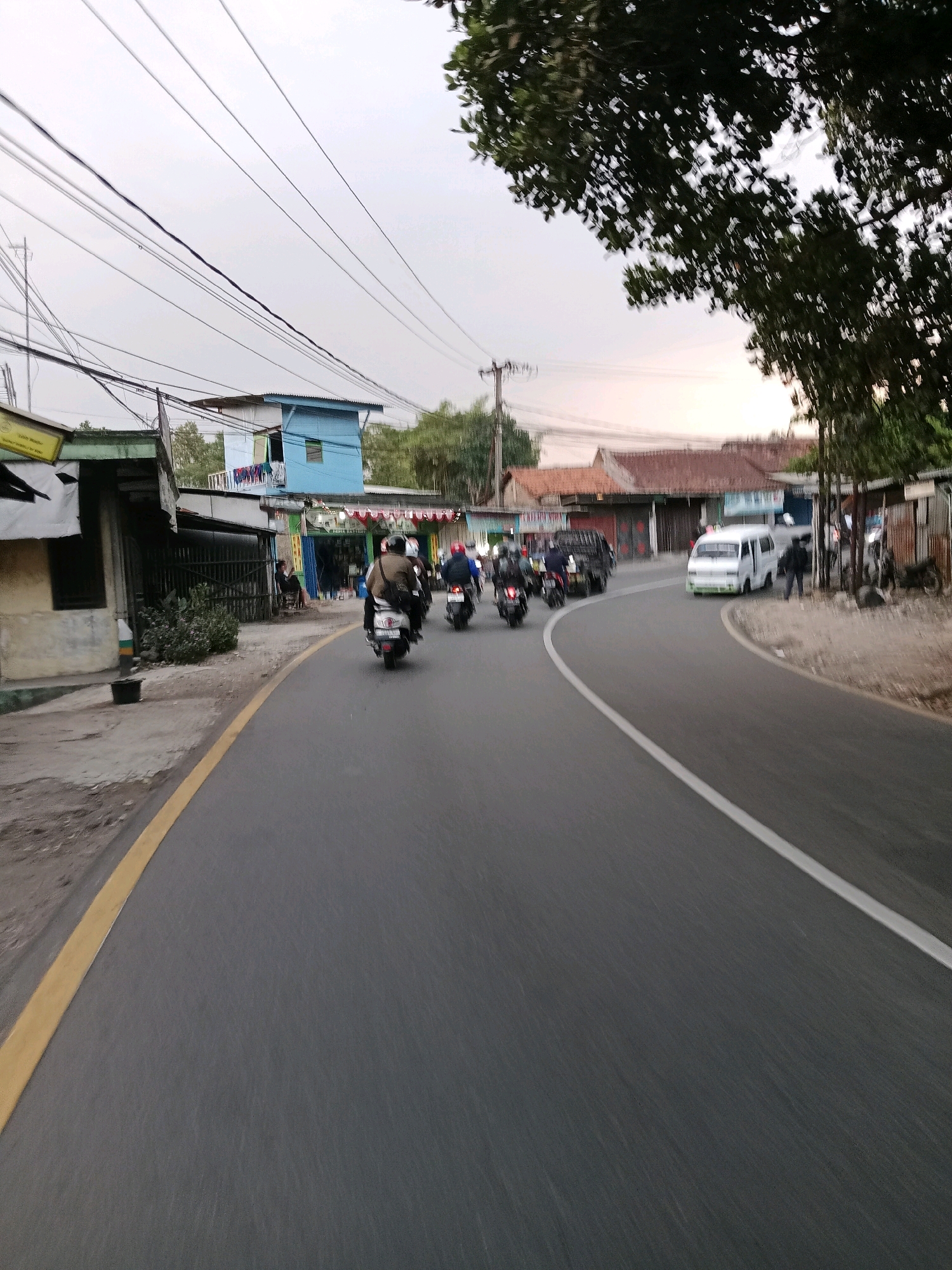 The image depicts a street scene in Sirnagalih, Indonesia, likely during the early morning or late afternoon due to the soft, diffused light and the absence of harsh shadows. The road, asphalted and marked with a yellow line on the left and a white line on the right, curves gently towards the right. A group of motorcyclists is traveling on the road, with the closest one, a scooter, moving away from the viewer. Further down the road, a white van is visible, and beyond that, more motorcyclists.
The environment is characterized by a mix of residential and commercial structures lining the street. On the left, there are low-rise buildings with visible signage and what appears to be a utility pole with numerous electrical wires crisscrossing overhead. A blue building with multiple stories stands out among the other structures. On the right side of the road, there are more buildings, some with corrugated metal roofs, and vegetation, including a large tree with dense foliage in the upper right corner. The sky is overcast, contributing to the overall muted lighting conditions. The scene suggests a typical day in a populated area, with ongoing traffic and daily activities.