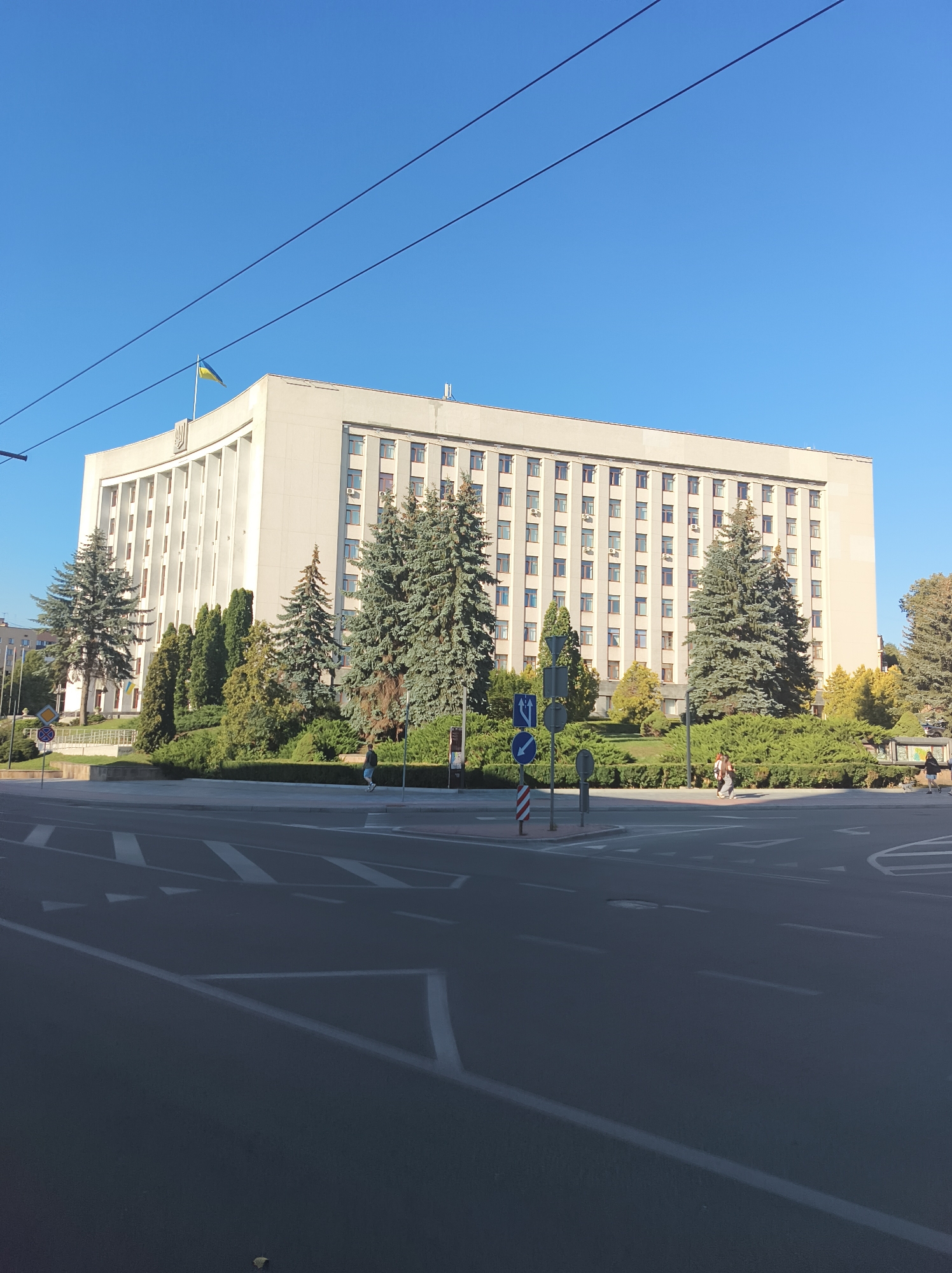 A wide, horizontal, outdoor, eye-level, medium shot captures the facade of a large, light-colored, institutional building in Ternopil, Ukraine. The building occupies much of the frame, with a clear, bright blue sky above. The building's architecture is characterized by a repetitive pattern of windows and what appear to be vertical architectural elements. A Ukrainian flag is visible flying from the top left corner of the building, near a central crest. In front of the building, a well-maintained green space with manicured bushes and trees is present. A paved road with lane markings is in the foreground, suggesting a city intersection. Several traffic signs are visible on the right side of the road. A few individuals are visible on the sidewalk in front of the building and crossing the street. The scene is bathed in natural daylight, indicating it was taken during the day.