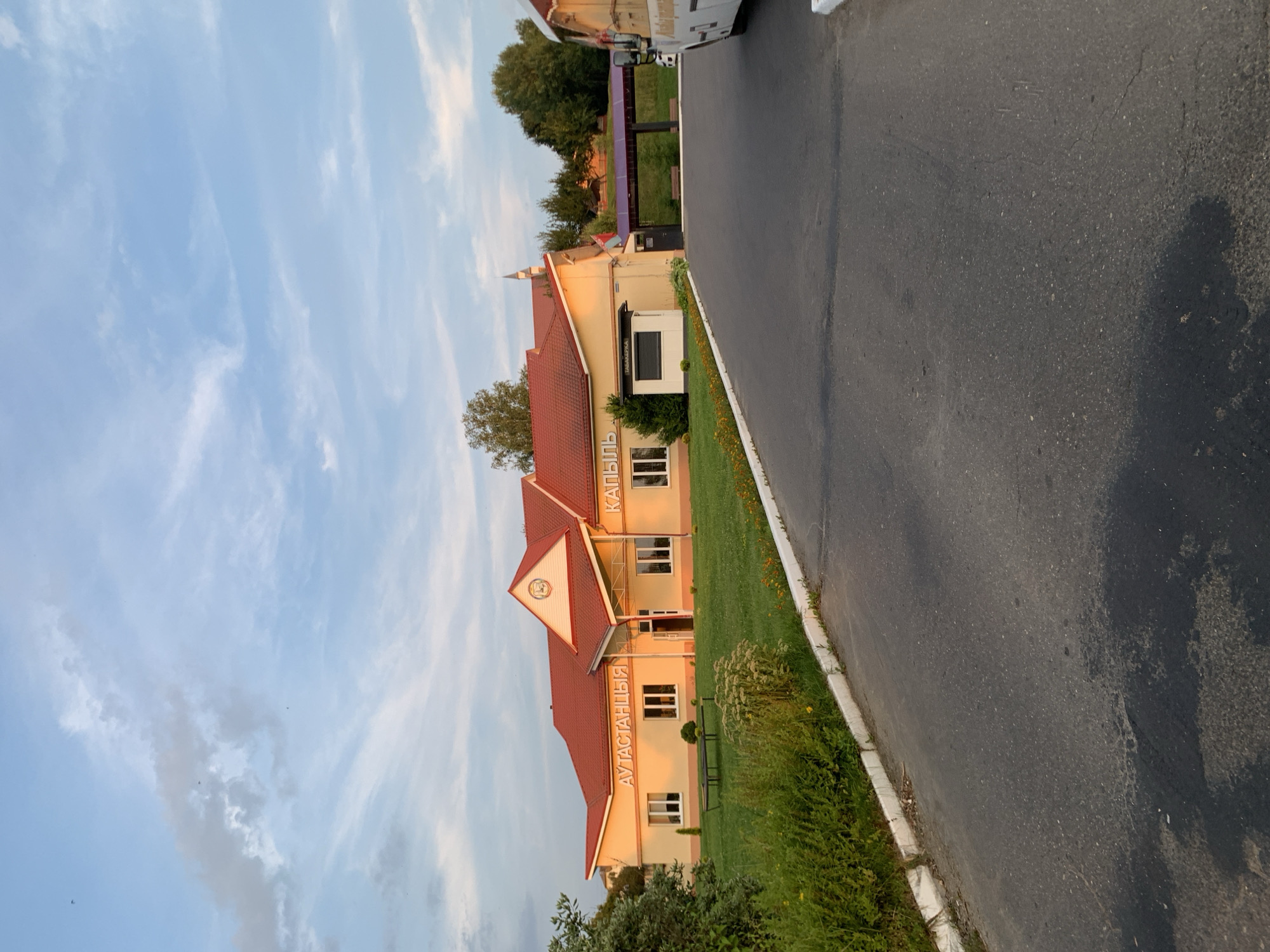 The image depicts a building with a red-tiled roof and yellow walls, situated in the city of Kapyĺ, Belarus. The building has multiple windows and is adorned with signage that reads "АУТАСТАНЦЫЯ" and "КАПЫЛЬ". In front of the building, there is a well-maintained green lawn with some sparse vegetation and a small, dark-colored bench. To the right of the building, a covered area with a purple roof is visible, and further to the right, the rear of a white bus is partially visible. The scene is set against a backdrop of trees and a sky with scattered clouds. The overall impression is of a public building or service center in a semi-rural or suburban setting.