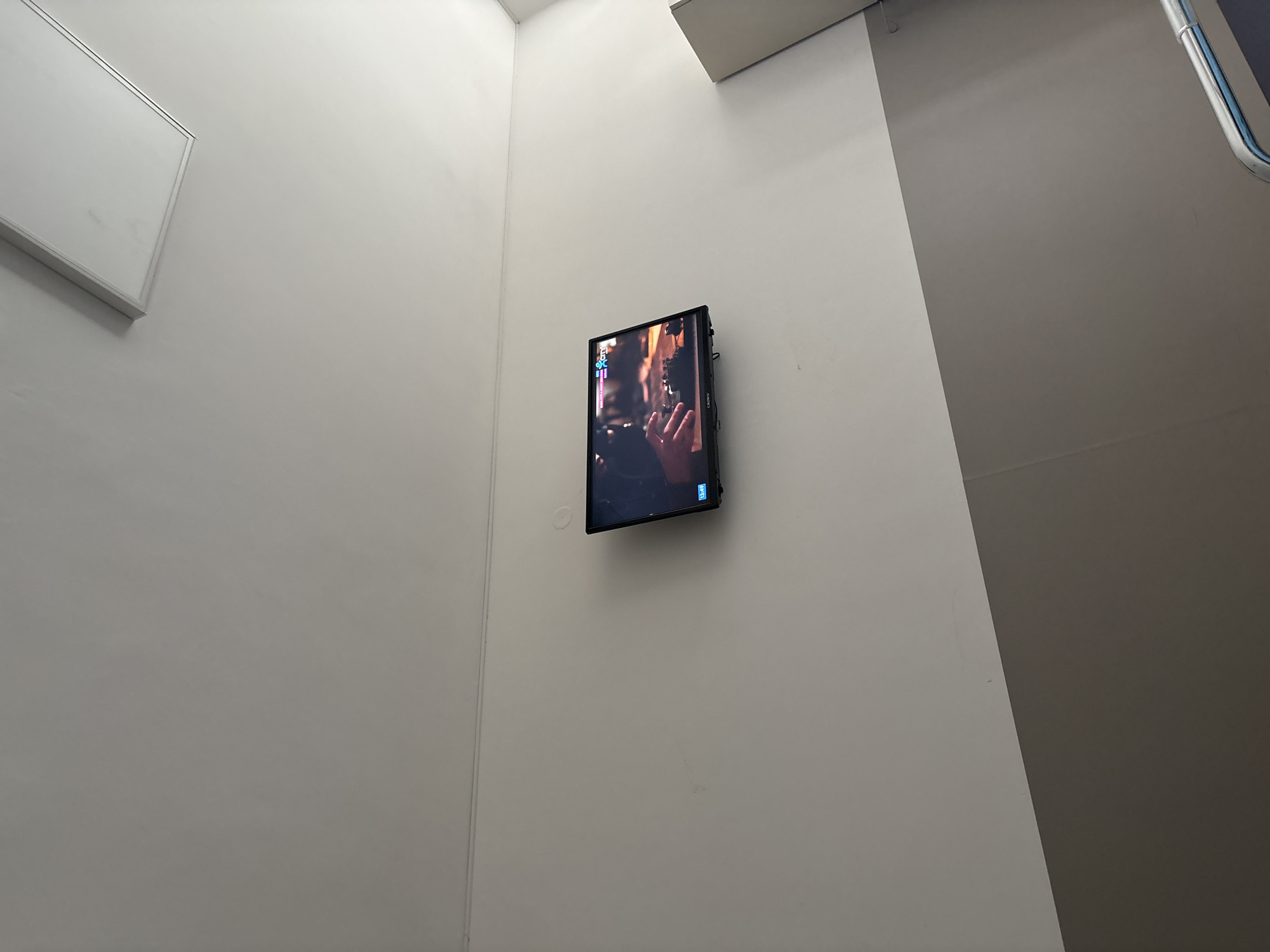 A wall-mounted flat-screen television displays a scene with a person's hand near a microphone, likely in an indoor setting. The television is centered on a white wall near the corner of the room. The location is an interior space, potentially in a building within Sofia, Bulgaria. There's a whiteboard on the left side and a corner with a grey wall on the right. A pipe is visible on the top right of the wall.