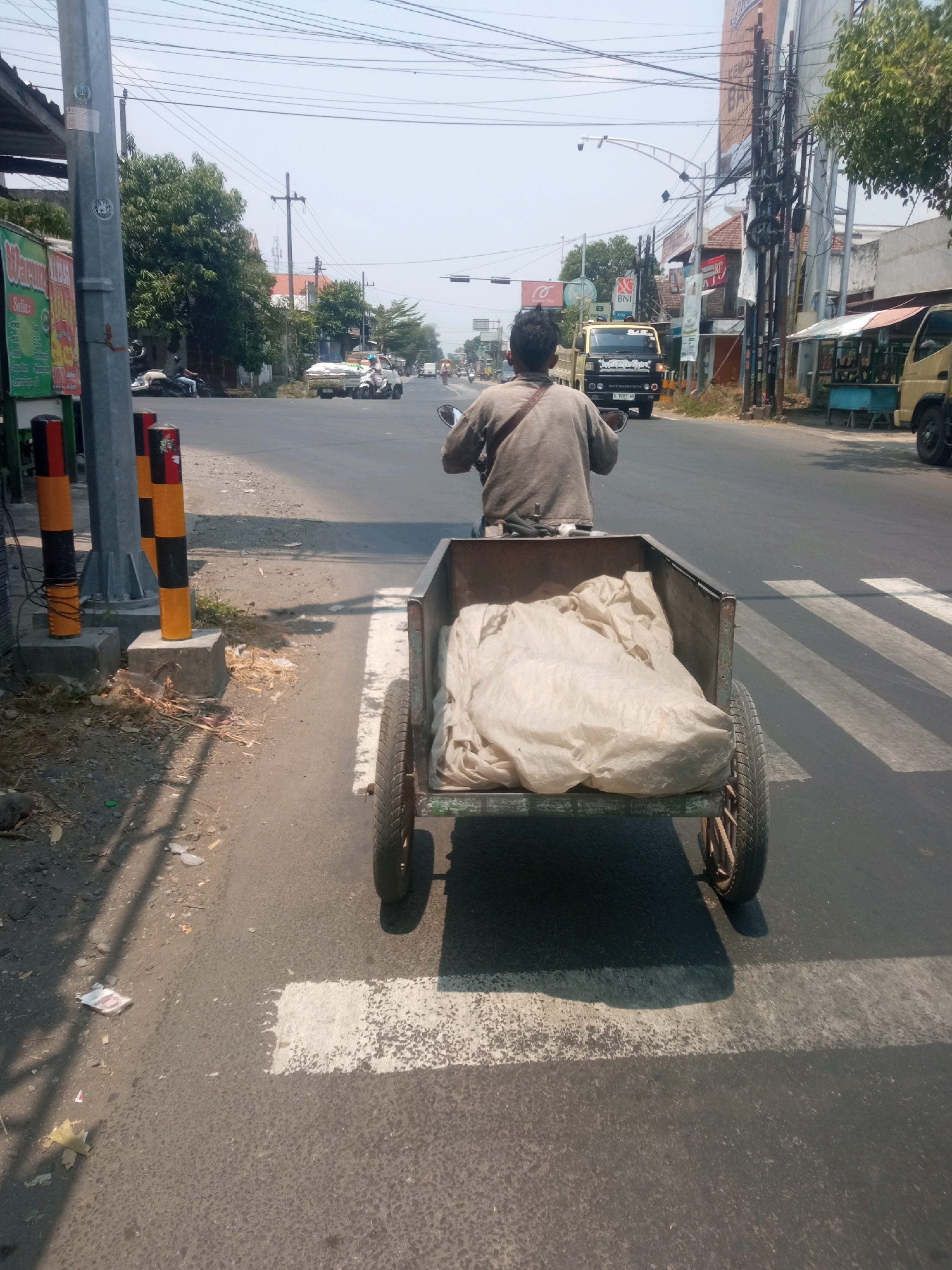 The image depicts a man riding a motorcycle with a trailer attached, moving through a street in Blimbing, Indonesia. The man, seen from behind, is wearing a neutral-colored shirt. The trailer is filled with a large, light-colored object wrapped in fabric. The motorcycle and trailer are on a paved road with a pedestrian crossing marked in white stripes. The surrounding environment includes buildings with awnings, utility poles with numerous wires, trees, and other vehicles, including trucks and cars, visible in the distance. The scene appears to be during the daytime, with bright sunlight casting shadows on the road.