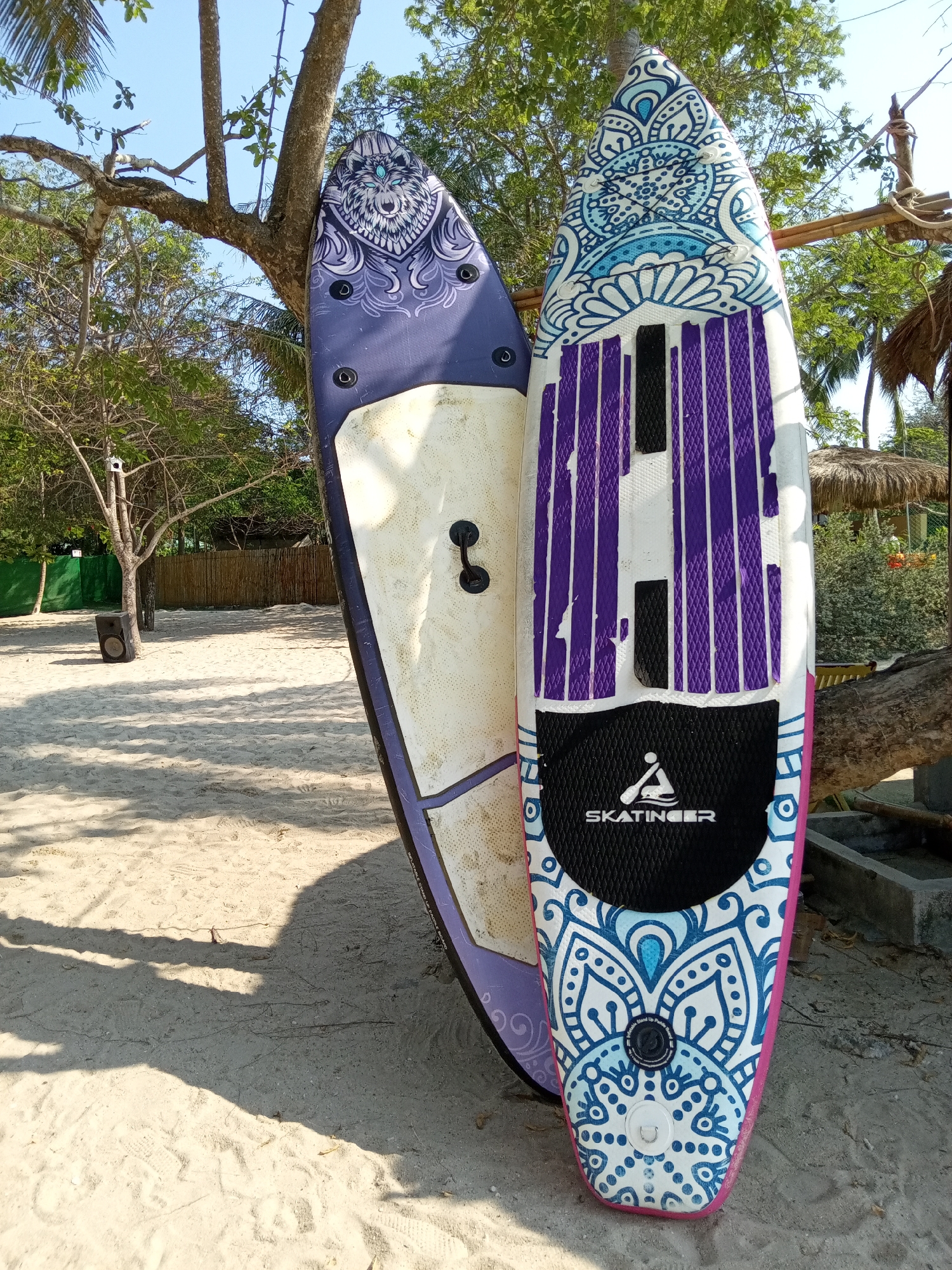 The image depicts two stand-up paddleboards (SUPs) leaning against a tree on a sandy beach. The paddleboard on the left is primarily purple, featuring a graphic of a wolf's head on its upper portion, while its underside is white. The paddleboard on the right has a decorative blue and white pattern with purple grip pads. A black section with the text "SKATINGER" is present on the lower portion of the right SUP. The background includes trees, a bamboo fence, a small structure with a thatched roof, and beach sand. The location is likely Nha Trang, Vietnam, based on the tropical vegetation and beach setting. A black speaker is located a few feet away.