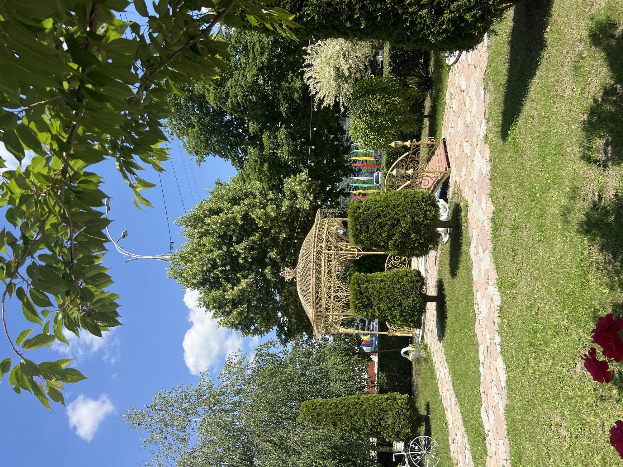 The image depicts a picturesque park scene, likely within the Dubăsari town council in Moldova, on a bright, sunny day. The focal point is a decorative, golden gazebo with intricate wrought-iron details, positioned centrally amidst lush greenery. A small, golden bridge with ornate railings and lampposts is situated to the right of the gazebo, spanning over what appears to be a dry stream bed or a decorative ditch.
The park features well-maintained pathways made of interlocking stone pavers in shades of beige and pink, winding through vibrant green grass. Several manicured evergreen trees are strategically placed throughout the landscape, adding structure and color. In the background, a variety of deciduous trees form a dense canopy, suggesting a mature park. Beyond the trees, colorful flags are visible, hinting at a festive or public event. A white decorative bicycle with a basket is visible on the left side of the image, adding a whimsical touch.
The sky is a clear, bright blue, dotted with a few fluffy white clouds, indicative of good weather. The overall atmosphere is serene and inviting, suggesting a well-kept public space designed for relaxation and enjoyment. No people are prominently featured in the image, allowing the focus to remain on the landscape and its decorative elements.