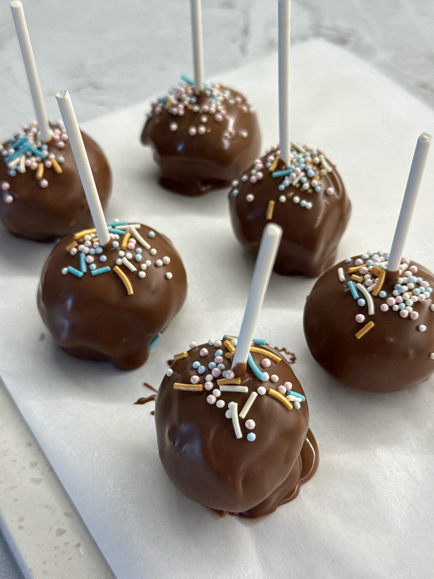 Six chocolate cake pops are arranged on a white surface. The cake pops are coated in dark chocolate and decorated with pastel-colored sprinkles, including blue, pink, and gold. Each cake pop is on a white stick. The background is a marble countertop. The scene is indoors. There is no information about the location in Lyubertsy, Russia.