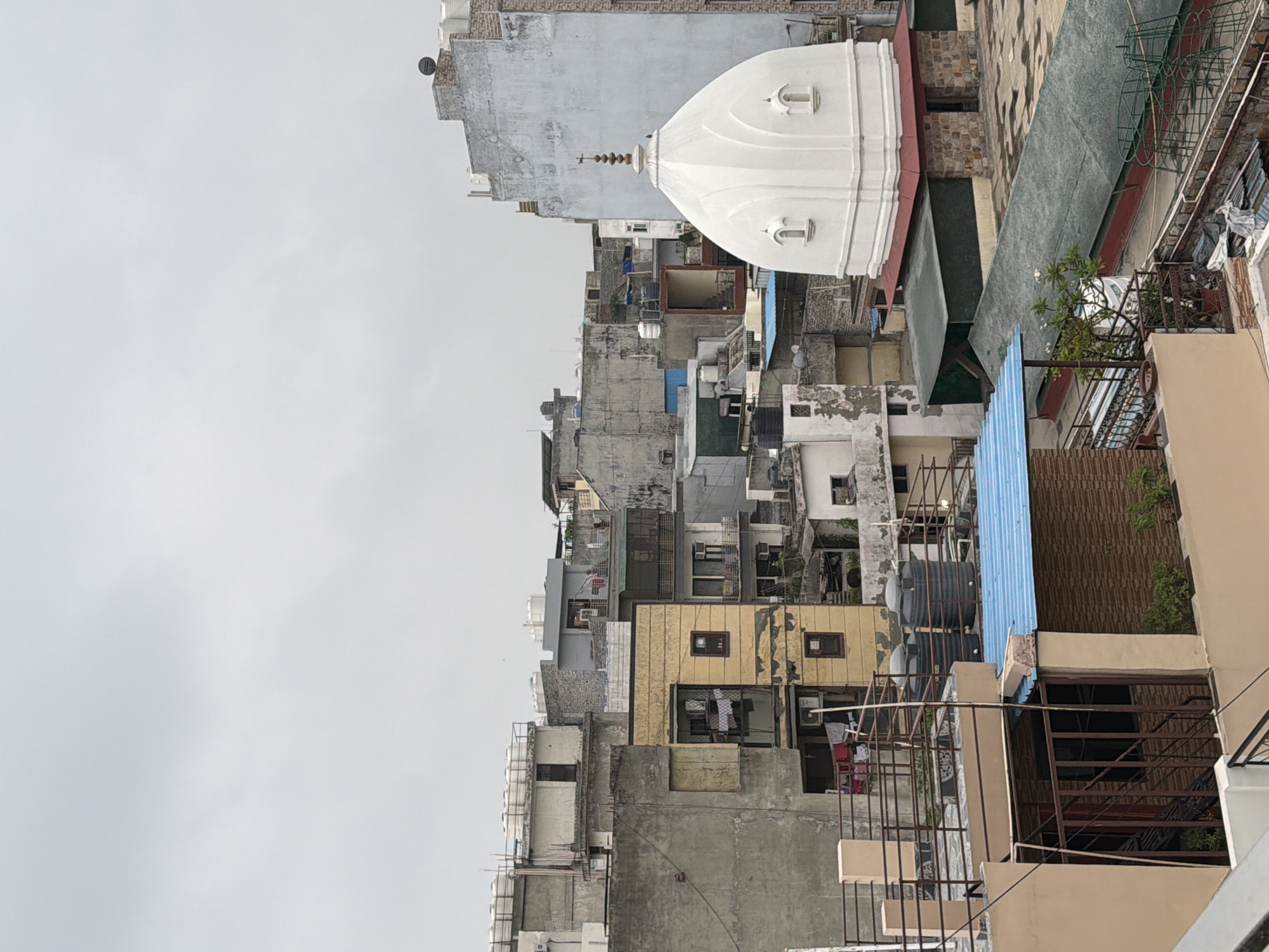 The image depicts an urban rooftop view in Delhi, India, characterized by densely packed buildings under an overcast sky. In the foreground, various structures with exposed water tanks on their roofs are visible, a common sight in Indian cities due to unreliable water supply. A white domed structure, possibly a temple or a part of a religious building, stands out against the muted tones of the surrounding architecture. The buildings themselves are a mix of styles and materials, with some appearing weathered and aged, while others show signs of recent construction or renovation. Laundry is visible hanging on clotheslines between buildings, suggesting residential use. The overall impression is one of a bustling, built-up urban environment typical of many Indian cities. There are no people visible in the frame.