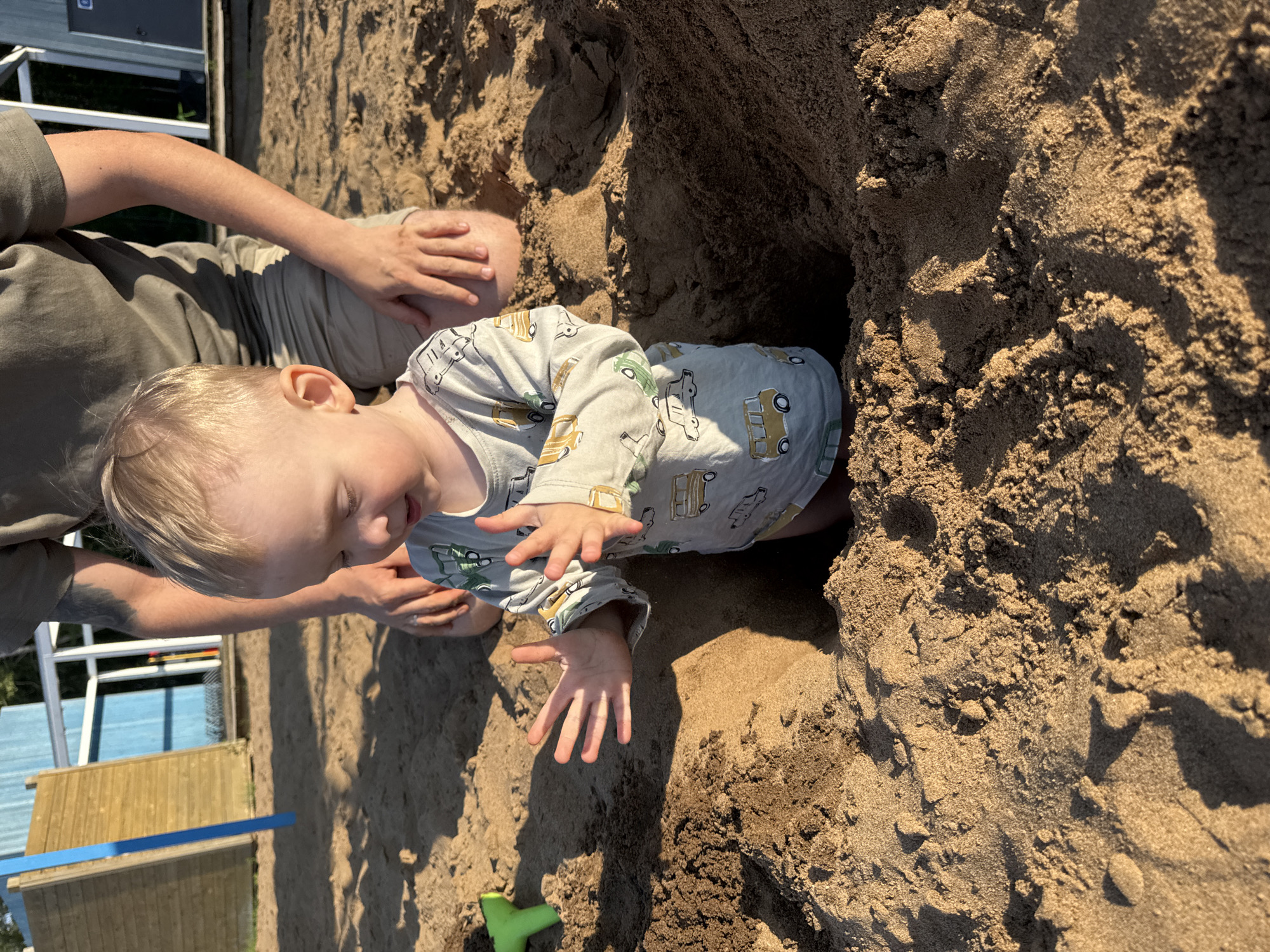 The image shows a close-up view of a young child playing in the sand. The child, with blonde hair, is wearing a light-colored long-sleeved shirt with a pattern of yellow and green vehicles. The child's hands are extended forward, appearing to be interacting with the sand. The child is partially submerged in a dug-out hole in the sand.
In the background, the arm and shoulder of an adult are visible, suggesting the adult is with the child and possibly assisting them. The adult is wearing a dark green t-shirt. The overall setting is a sandy area, likely a beach or a playground, given the presence of sand. There are also glimpses of what appear to be playground structures in the background, including a wooden structure with blue accents and a metal railing. The sunlight casts shadows on the sand, indicating it is daytime. The child's expression and posture suggest engagement and enjoyment in their activity.
The location context is Chaikovsky, Russia. The time of day appears to be sunny and warm, conducive to outdoor play. There is no visible text in the image.
