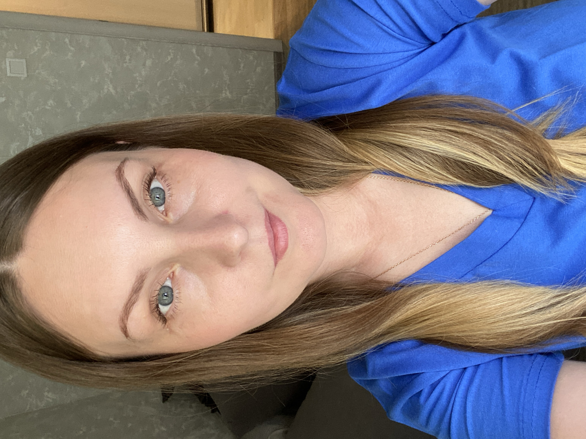 A selfie depicts a woman with long, light brown hair and blue eyes, wearing a blue t-shirt. A thin gold necklace is visible around her neck. The background consists of a grey wall with a faint pattern and part of a wooden cabinet. The location is unspecified, and there is no indication it is in Noviy, Russia.