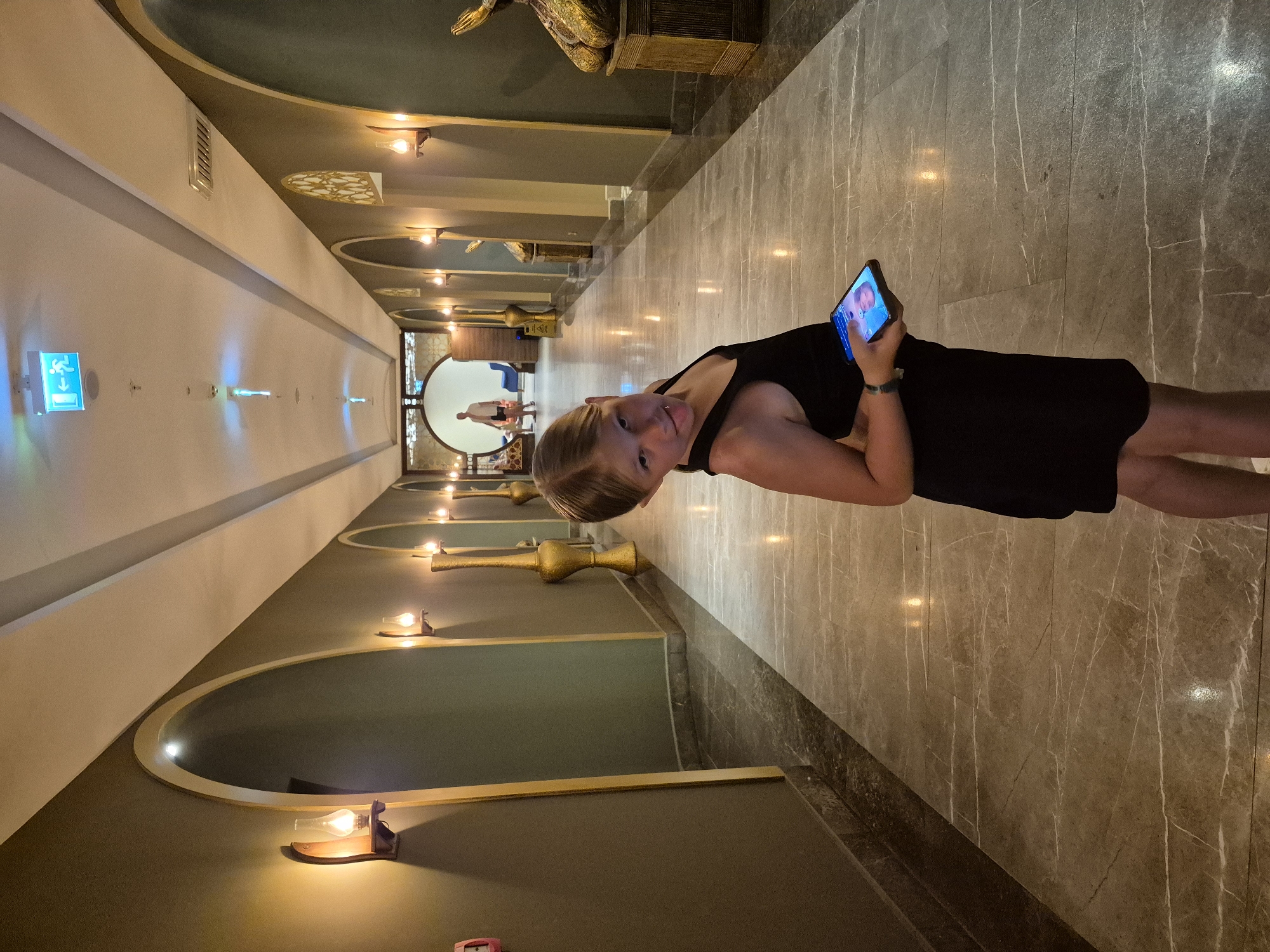 A young woman stands in a hotel hallway in Alanya, Turkey. She is holding a smartphone and looking at the screen. She is wearing a black dress and is positioned on a polished marble floor. The hallway is decorated with ornate lighting fixtures and arched doorways. An exit sign is visible in the distance.