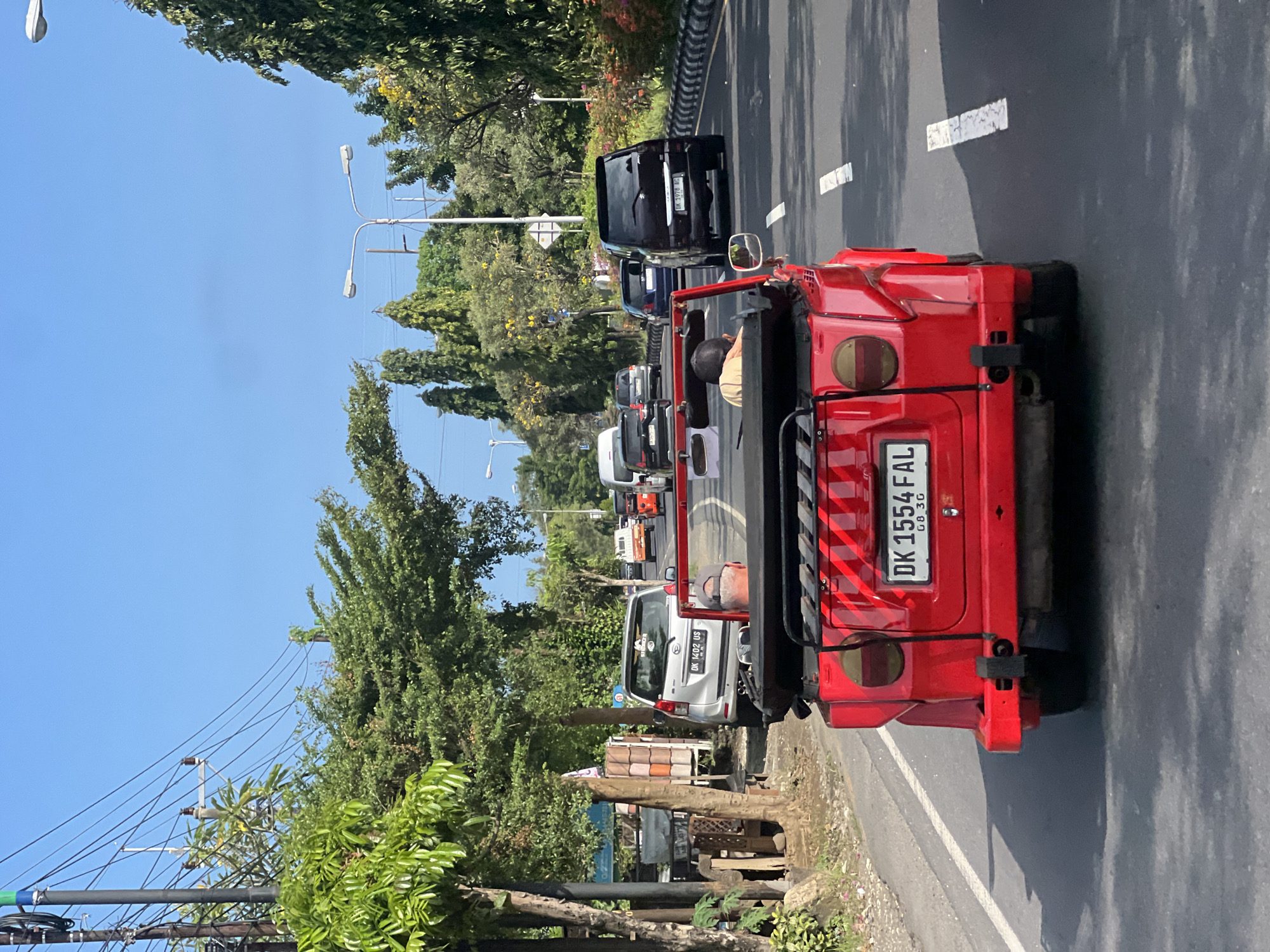 The image depicts a traffic scene in Denpasar, Indonesia. The focus is on the rear of a red vehicle, distinguished by a license plate "DK 1554 FAL". The vehicle is positioned on the right side of a multi-lane road, with other cars visible in the distance, indicating a moderate level of traffic. The background shows a mix of urban elements and nature, including trees, street lights, and power lines against a clear blue sky. On the left side of the road, there is a view of the sidewalk with wooden construction, plants and other cars.