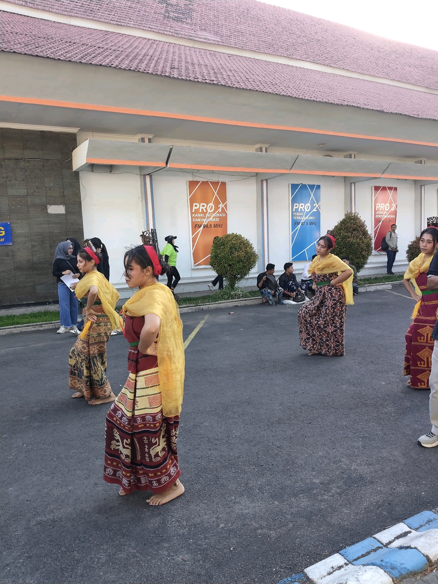 The image depicts an outdoor performance in front of a building in Malang, Indonesia. Several young women are dressed in traditional attire, appearing to be performing a dance. The attire consists of colorful, patterned skirts, sashes, and headbands. The women are barefoot. In the background, there are banners with the text "PRO X," "PRO 2," and "PRO 3," likely indicating a radio station or event. Several people are seated on the ground watching the performance, while others stand or walk by. The environment appears to be a paved outdoor area, possibly a parking lot, with a building facade behind the performers. The architecture of the building suggests a modern design with a tiled roof and large windows. The lighting indicates it is daytime.