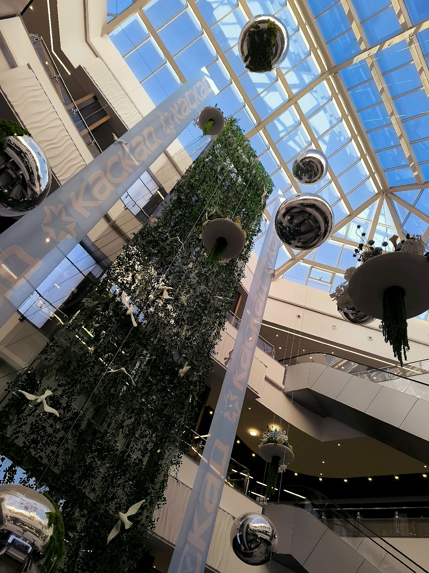 The image captures an interior view of a modern shopping mall in Qarağandı, Kazakhstan, focusing on its atrium and decorative elements. The scene is characterized by a multi-level structure with escalators and balconies visible in the background. A large, vertical green installation, resembling a plant-covered tower, dominates the central part of the frame. Hanging from the ceiling and around this installation are several large, reflective spherical ornaments and smaller decorative elements that appear to be hanging planters with foliage. White bird figurines are strategically placed among the greenery. A prominent, tall, white banner with blue lettering and a star logo, likely a brand advertisement, runs vertically through the composition. The ceiling is a glass-covered structure allowing natural light to filter into the space, revealing a clear blue sky above. The overall impression is one of a contemporary, well-lit commercial environment with a focus on aesthetic decoration and natural elements.