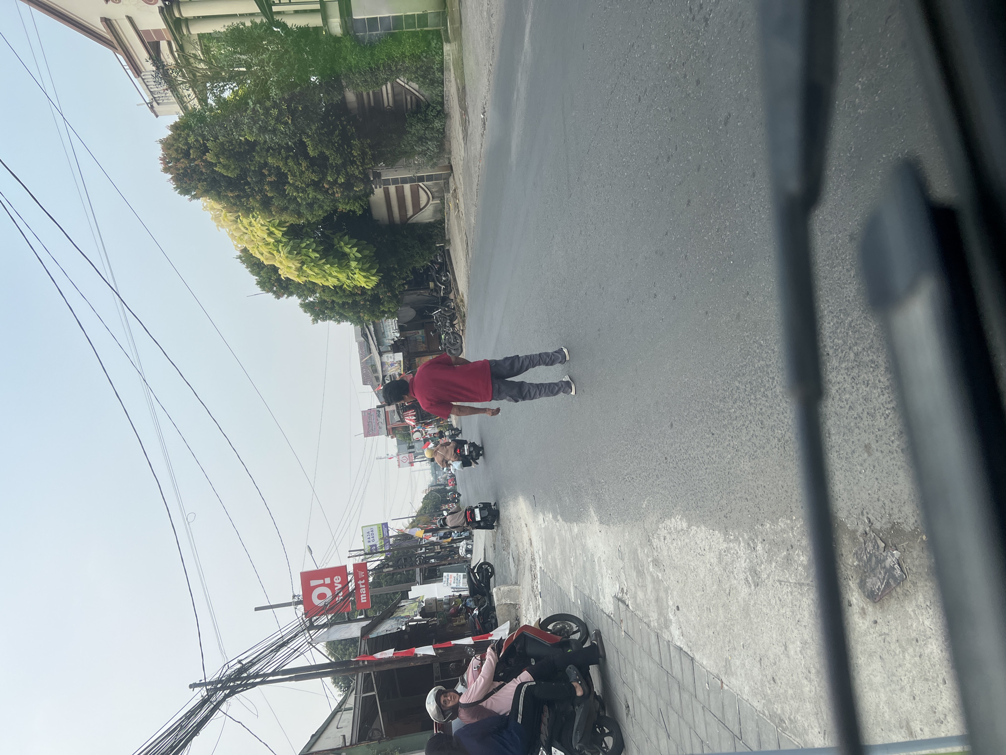 The image depicts a street scene in Citeureup, Indonesia, with several people on motorcycles. In the foreground, a person wearing a red shirt and gray pants stands in the middle of the road, facing away from the camera. To the left, two individuals are seated on a motorcycle parked on the sidewalk, with one person wearing a helmet and a pink shirt. Behind them, several other motorcycles are visible, some parked and some in motion. Buildings line the left side of the street, featuring signs, including one that reads "O! SAVE" and another with "mart". Trees and overhead power lines are also prominent features of the environment. The road surface is asphalt. The sky is overcast.