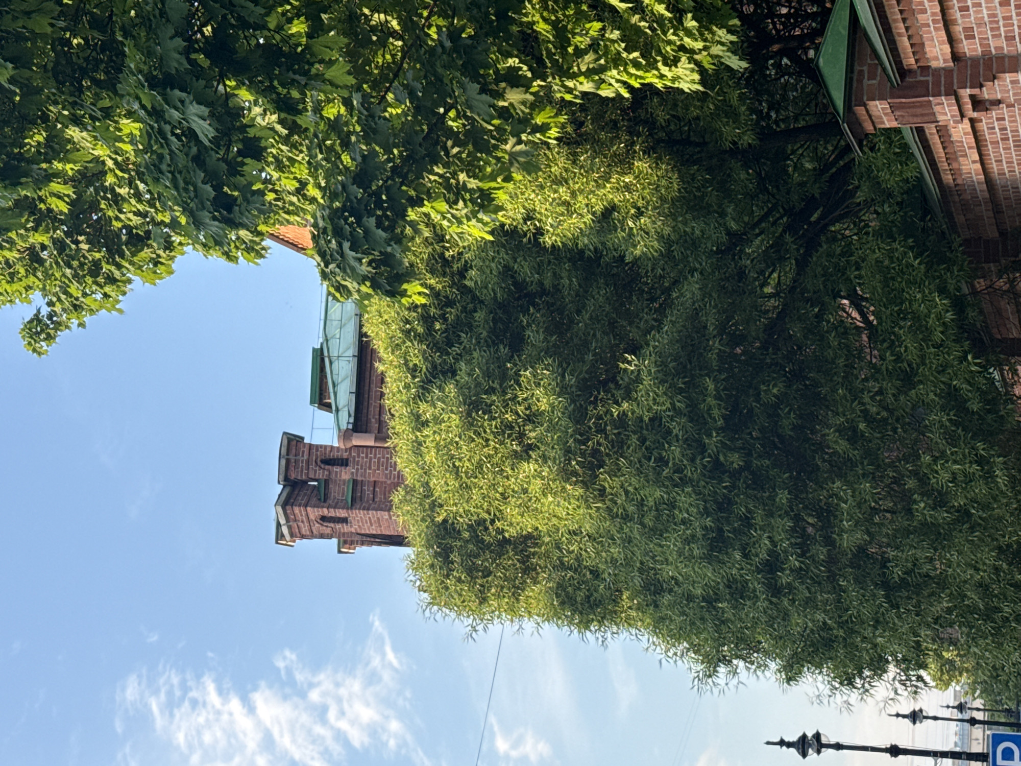 The image depicts a street scene in Saint Petersburg, Russia, under a bright, clear sky with scattered cirrus clouds. A red brick building, possibly a church or historical structure, is partially visible, framed by dense green foliage. The architecture includes tiered elements and what appears to be a copper or green-colored roof section. The foliage suggests mature trees along the street. At the bottom of the image, there are indications of street furniture or signage. The overall impression is a blend of urban architecture and natural greenery, typical of Saint Petersburg's cityscape.