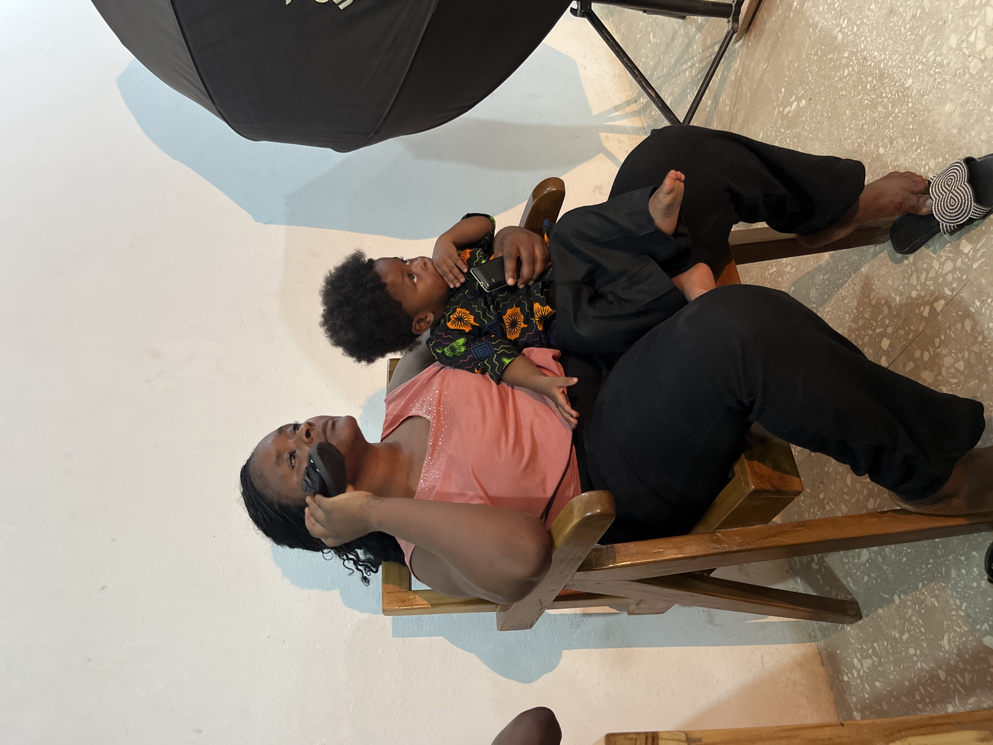 The image shows a woman and a child sitting on a wooden chair. The woman is wearing a pink sleeveless top with a beaded neckline and black pants. She has dark, braided hair and is looking up and to her right, holding a black object to her ear as if on a phone call. Her left hand is resting on her leg. The child is lying on the woman's lap, facing away from the viewer, and is wearing a black shirt with an orange and green floral pattern and black pants. The child's left hand is near their face, and their feet are visible.
The setting appears to be indoors, with a white wall and a speckled floor. A large, black umbrella-like object, possibly a photography light modifier, is visible in the upper left corner of the frame, casting a shadow on the wall. The lighting suggests it might be daytime. The overall mood is relaxed and intimate, with the child appearing comfortable on the woman's lap. No visible text is present in the image.