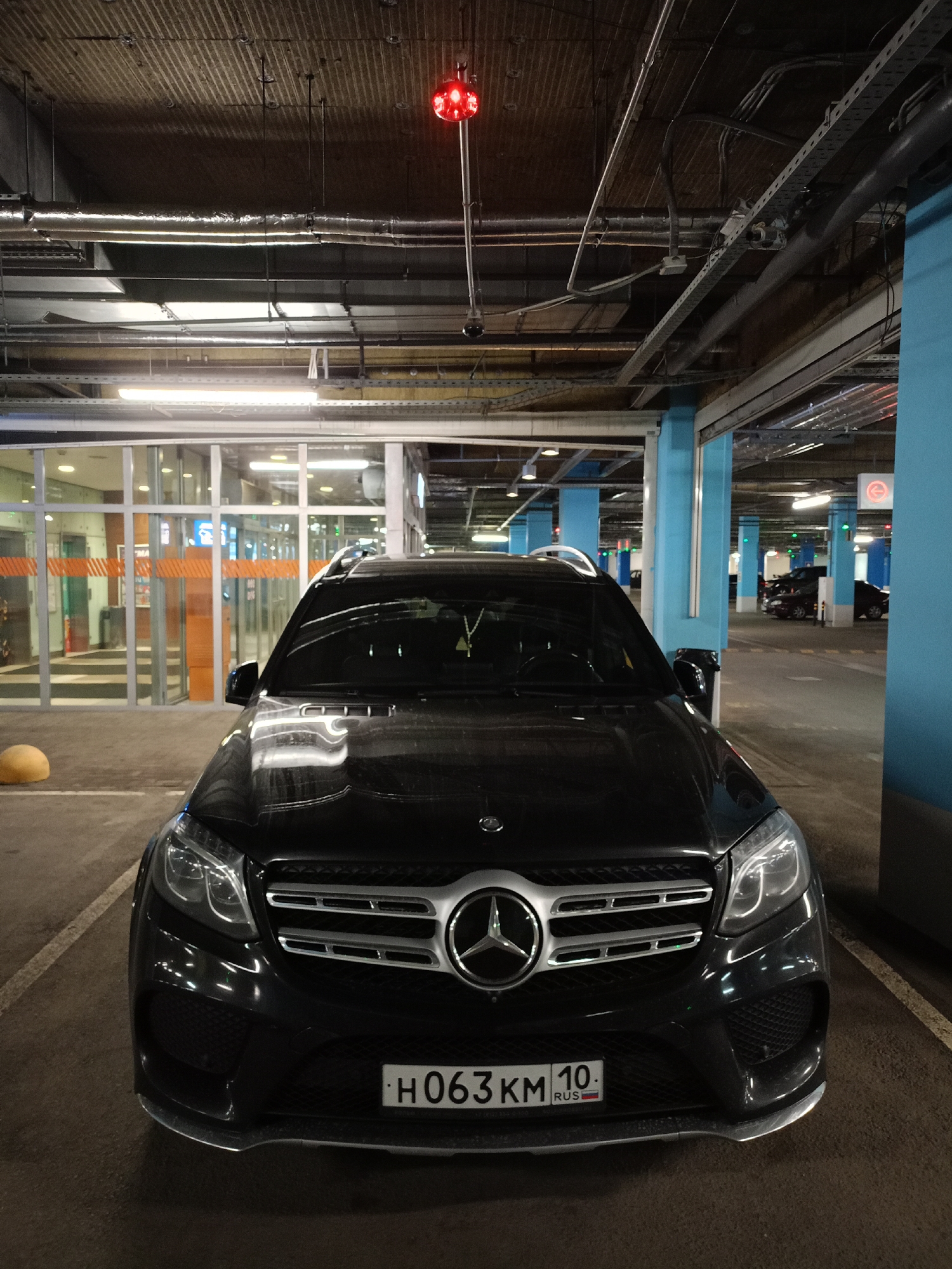 The image shows a black Mercedes-Benz SUV parked in an indoor parking garage. The car is the primary subject, positioned centrally in the frame, facing the viewer. It has its headlights on. The license plate reads "H063KM 10 RUS".
The setting is a dimly lit parking garage with concrete pillars painted blue and a corrugated metal ceiling. Exposed pipes and wiring are visible on the ceiling. Fluorescent lights illuminate the space, and a red traffic signal is suspended from the ceiling above the car. In the background, a glass-fronted building or entrance area is visible, with orange-striped barriers. Another car is parked in the distance. The ground is asphalt, marked with parking lines. A yellow spherical object, possibly a bollard, is partially visible on the left side of the frame. The overall impression is of a typical urban parking facility, likely at night or in low light conditions. There are no people visible in the image.