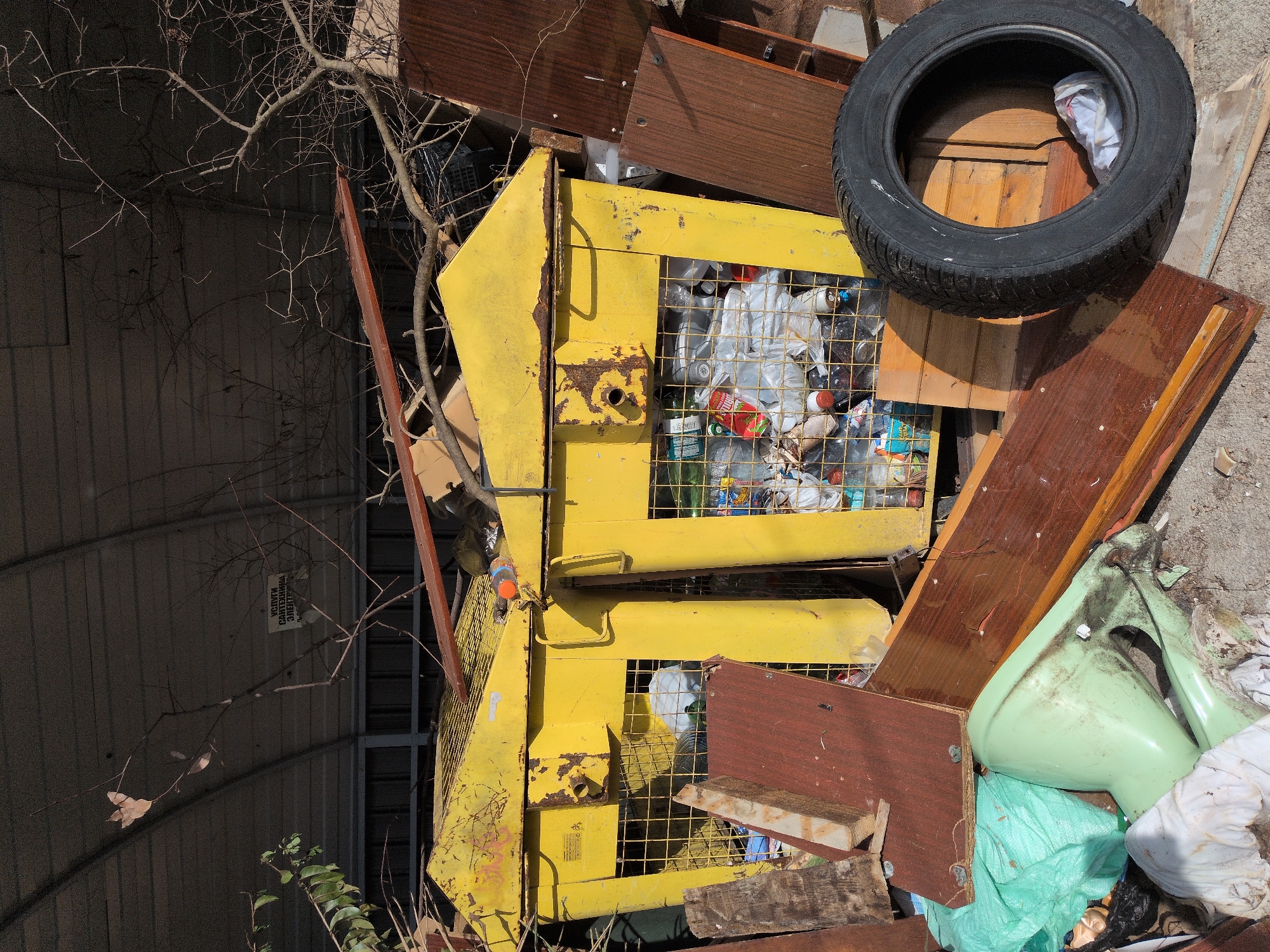 This image captures a cluttered scene of discarded items and waste, likely at a makeshift dump or recycling collection point in Sochi, Russia.
In the foreground and central to the image are two large, yellow metal bins or cages, heavily rusted and showing signs of wear. The bins are filled with various types of refuse, predominantly plastic bottles and plastic bags, indicating they might be intended for recycling or general waste collection.
Surrounding and resting upon these bins and the ground are numerous other discarded objects. Several dark brown wooden planks and boards are scattered around, some appearing to be parts of old furniture. A prominent black car tire rests on top of some wooden pieces and partially on one of the yellow bins in the upper right. Near the bottom right, a broken, light green ceramic object, resembling a toilet bowl or sink base, is visible, surrounded by a light green plastic bag and other debris.
Bare tree branches, largely devoid of leaves, extend from the left side across the scene, casting shadows. In the background, behind the branches, is a dark corrugated metal structure, possibly a wall or part of a building. The ground appears to be a mix of dirt and possibly broken concrete.
The scene is brightly lit by natural sunlight, suggesting it is daytime, with clear shadows visible. The overall impression is one of neglect and environmental disarray.
Visible text includes a piece of paper or cardboard hanging from a bare branch in the upper left background, which displays Cyrillic text: "УСЛУГИ САНТЕХНИКА ЭЛЕКТРИКА..." (Services: Plumber, Electrician...). A small, unreadable label or sticker is also present on the side of the lower yellow bin.