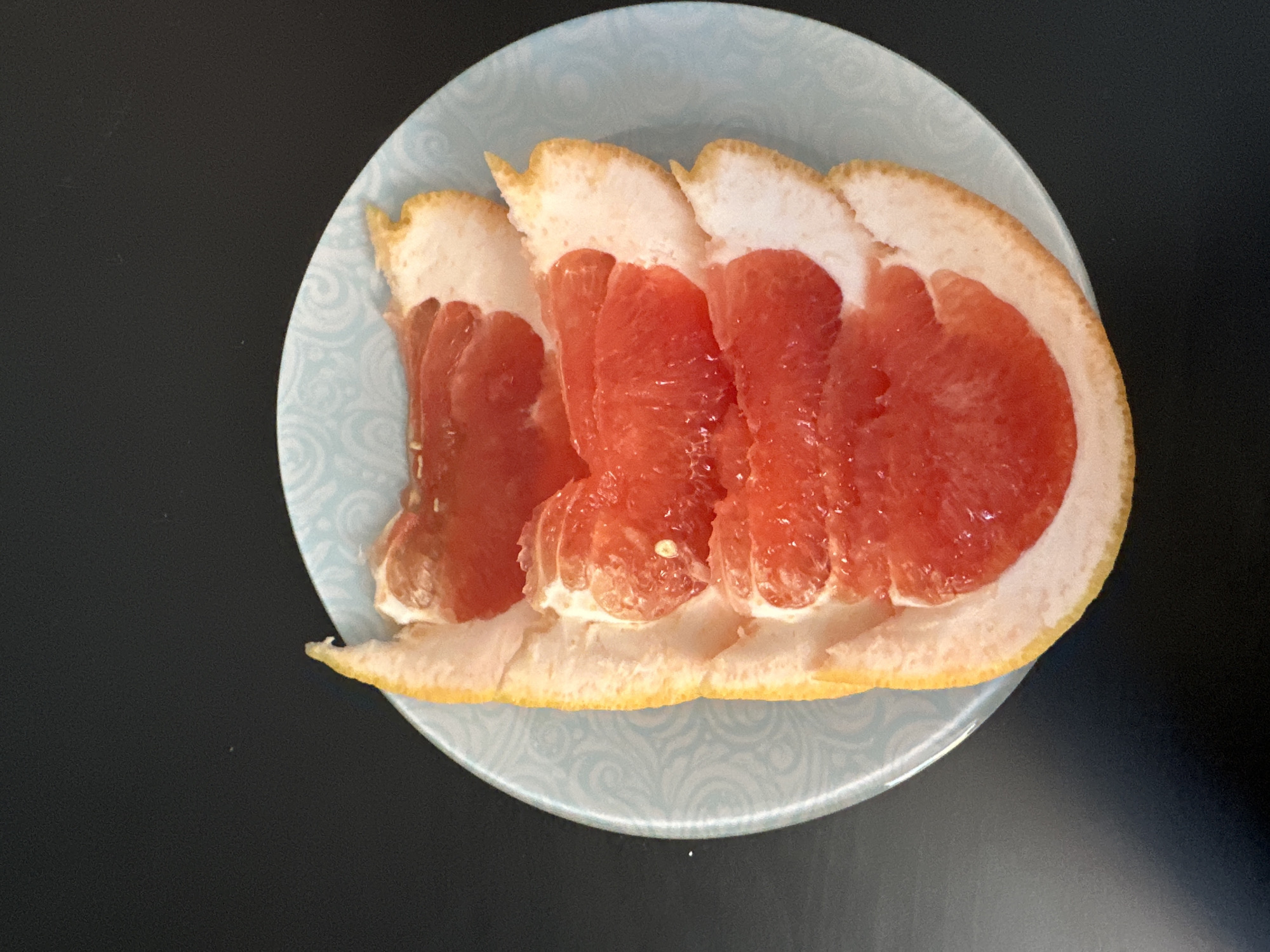 A close-up shot shows a peeled and segmented pomelo fruit served on a light blue plate with a swirling pattern. The pomelo's segments are a vibrant red, with the thick white pith still attached to the fruit. The plate rests on a dark gray surface. There is no indication of location.