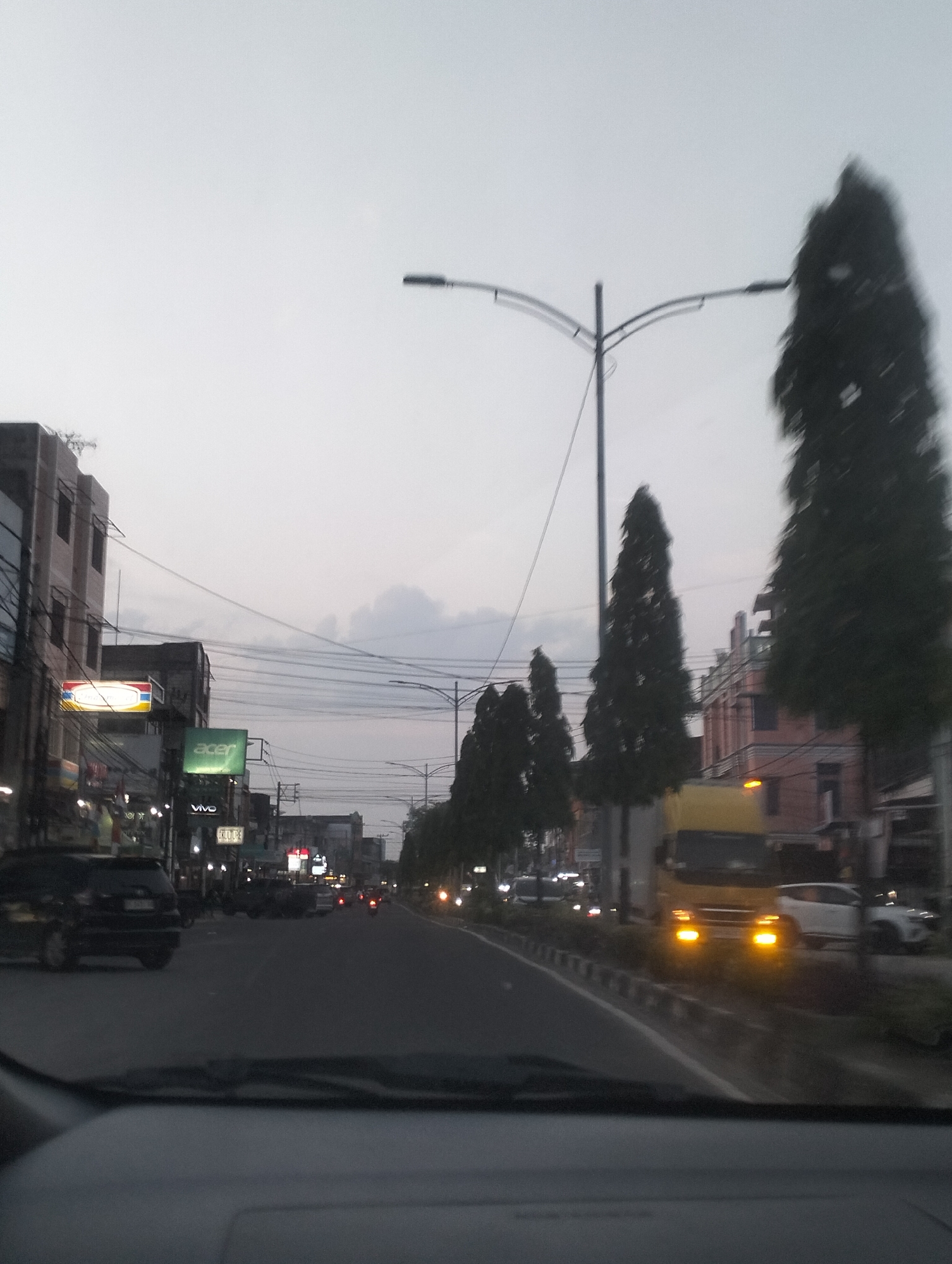 The image captures a street scene in Dumai City, Indonesia, likely during twilight. The perspective is from inside a vehicle, looking forward through the windshield. A paved road dominates the foreground, with vehicles including cars and a yellow truck visible. Streetlights are present, and storefronts with illuminated signs line the left side of the road. Trees are interspersed along the street, some appearing tall and slender. The sky is overcast with a pale, muted light. The overall impression is of a typical urban environment at dusk, with ongoing traffic and commercial activity.