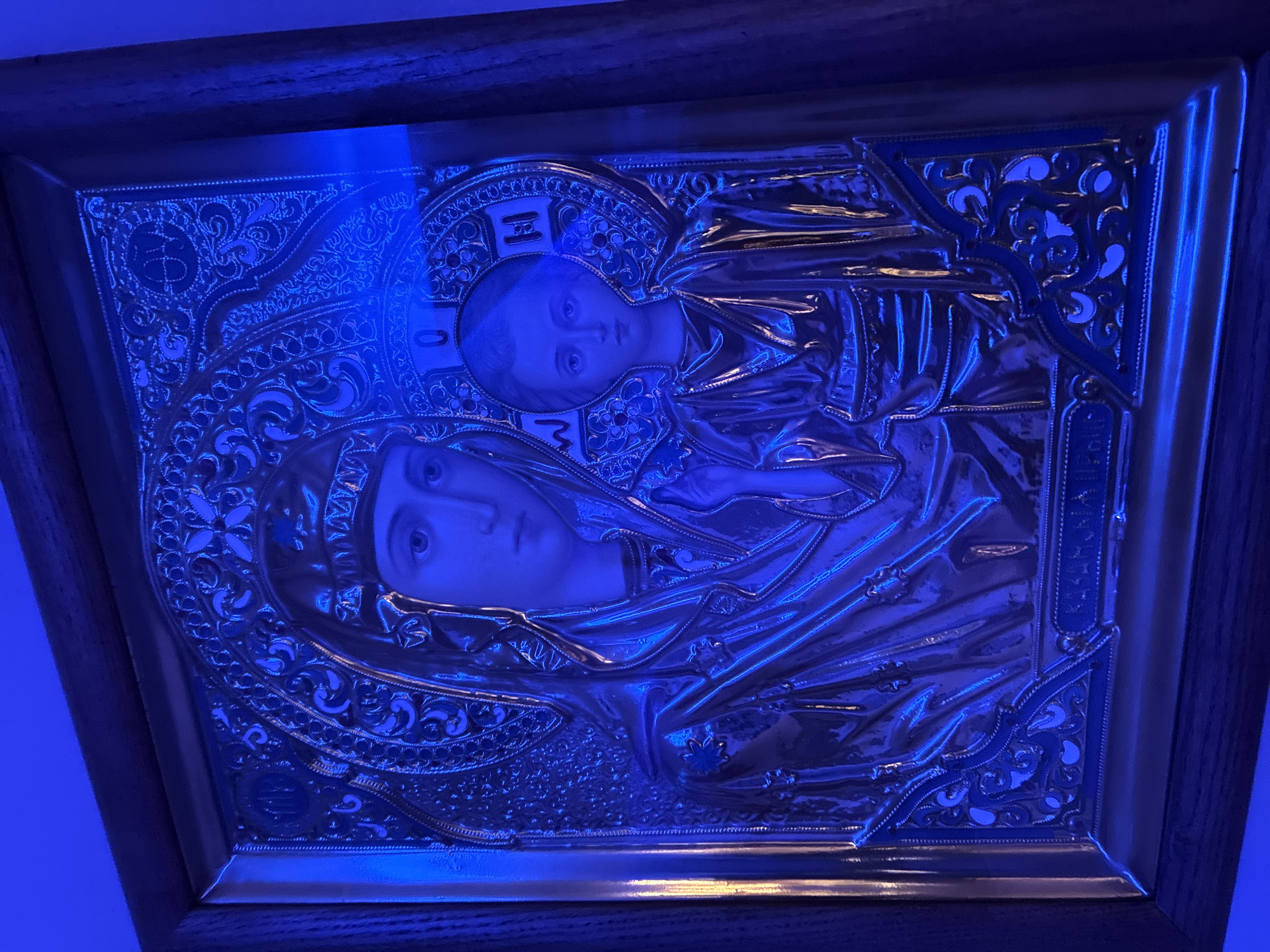 An ornate, silver-toned icon depicts the Virgin Mary holding the Christ child. Both figures are rendered in relief with detailed facial features and flowing garments. The Virgin Mary is on the left, her head slightly bowed and her gaze directed downward. The Christ child is on the right, looking directly at the viewer with a serious expression. Both figures are encircled by intricate decorative elements within a halo. The icon is framed by a dark wooden border. The entire scene is illuminated by a strong blue light, casting shadows and highlighting the metallic sheen of the icon. The background appears to be a wall, also bathed in blue light.