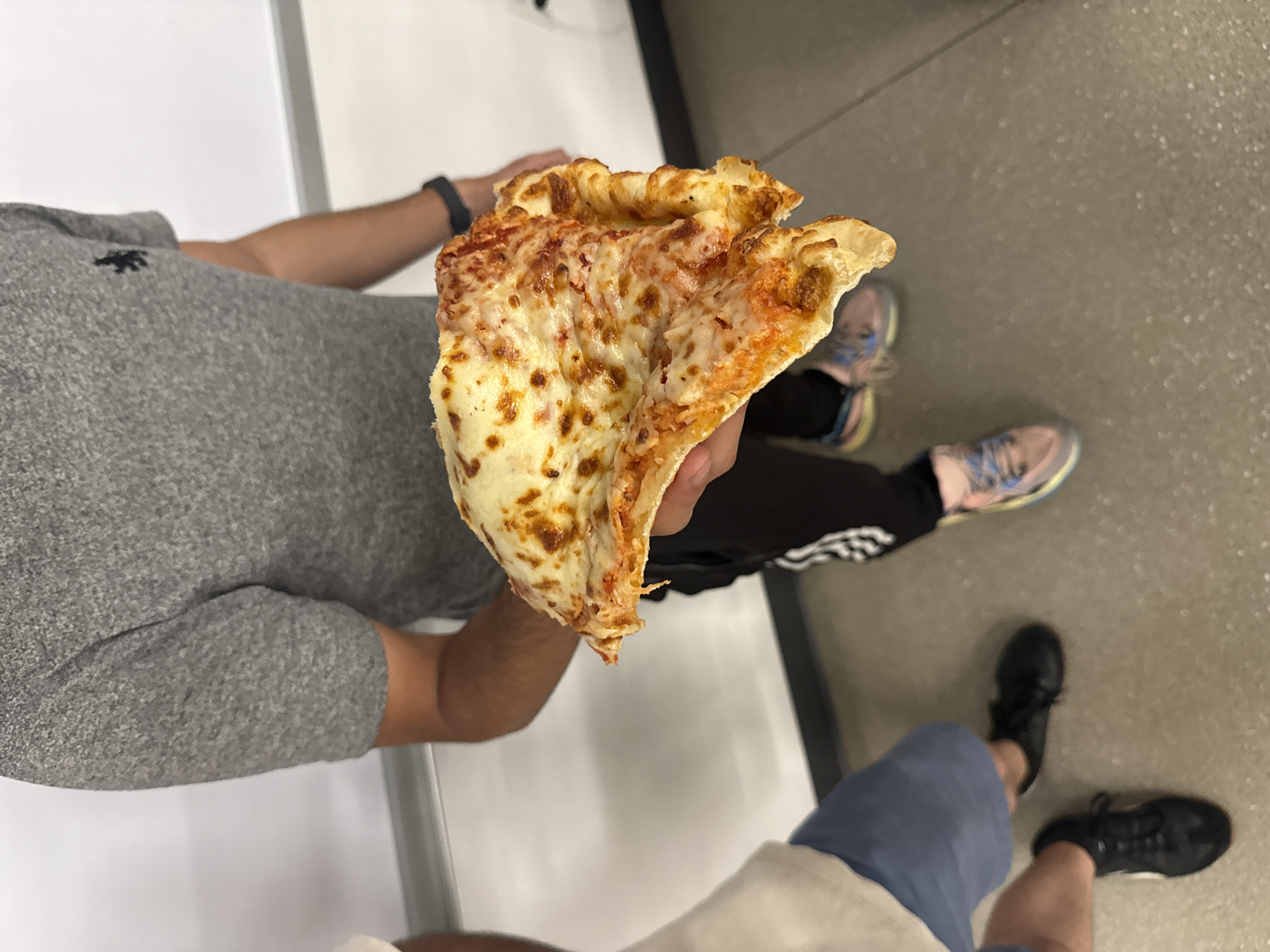 An overhead shot captures two individuals in an indoor setting, likely a casual public or commercial space in Mexico City, Mexico. The main subject is seen from the torso down, holding a large, folded slice of cheese pizza. This person wears a grey short-sleeved shirt with a small, dark embroidered logo (possibly an animal silhouette) on the chest, a black smartwatch on their left wrist, black pants with white stripes (reminiscent of Adidas apparel), and light pink and blue athletic sneakers.
The pizza slice is a classic cheese variety, with melted, browned cheese and visible red sauce, folded in half as if ready for a bite. Partially visible in the lower right corner are the legs and feet of a second person, wearing blue shorts and black sneakers. The floor is a neutral grey, and the background consists of plain, light-colored walls, suggesting an unadorned, functional environment like a food court, cafeteria, or large retail store. The lighting indicates an indoor setting. No explicit text is clearly visible apart from the ambiguous shirt logo and potential branding on the pants. The scene depicts a casual moment, possibly during a meal or break.