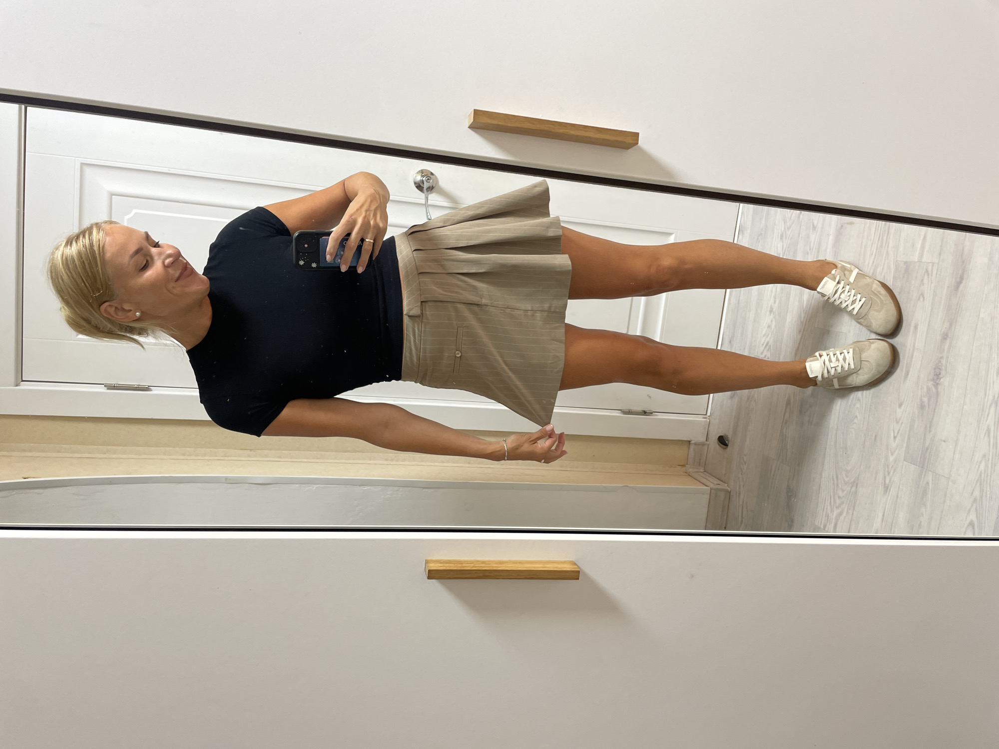 A blonde woman stands in front of a full-length mirror, taking a selfie. She is wearing a black t-shirt, a tan pleated mini-skirt with pinstripes, and white sneakers. Her hair is pulled back in a ponytail. The background appears to be an indoor setting, possibly a bedroom or hallway, with light-colored wood flooring and a white door. There are two wooden handles on a white closet door to the left of the frame. The mirror reflects the woman and her surroundings.