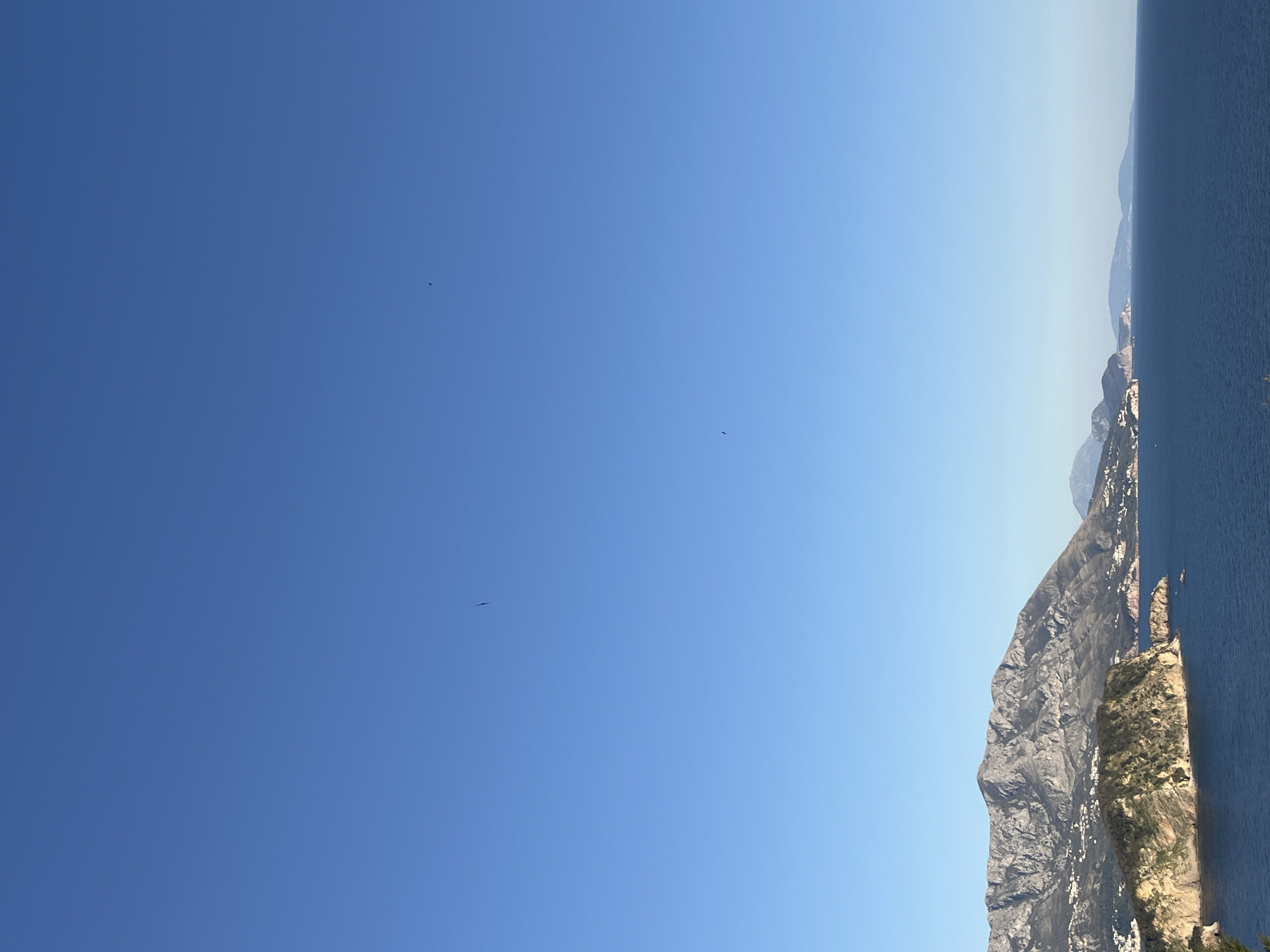 The image shows a landscape in Budva, Montenegro. The right side features a rugged, mountainous coastline meeting the Adriatic Sea, with visible cliffs and vegetation. The left side is dominated by a clear blue sky, with three small birds visible in the distance. The photo appears to be taken during the day, with good visibility. The sky transitions from a darker blue on the left to a lighter blue toward the horizon where it meets the sea.