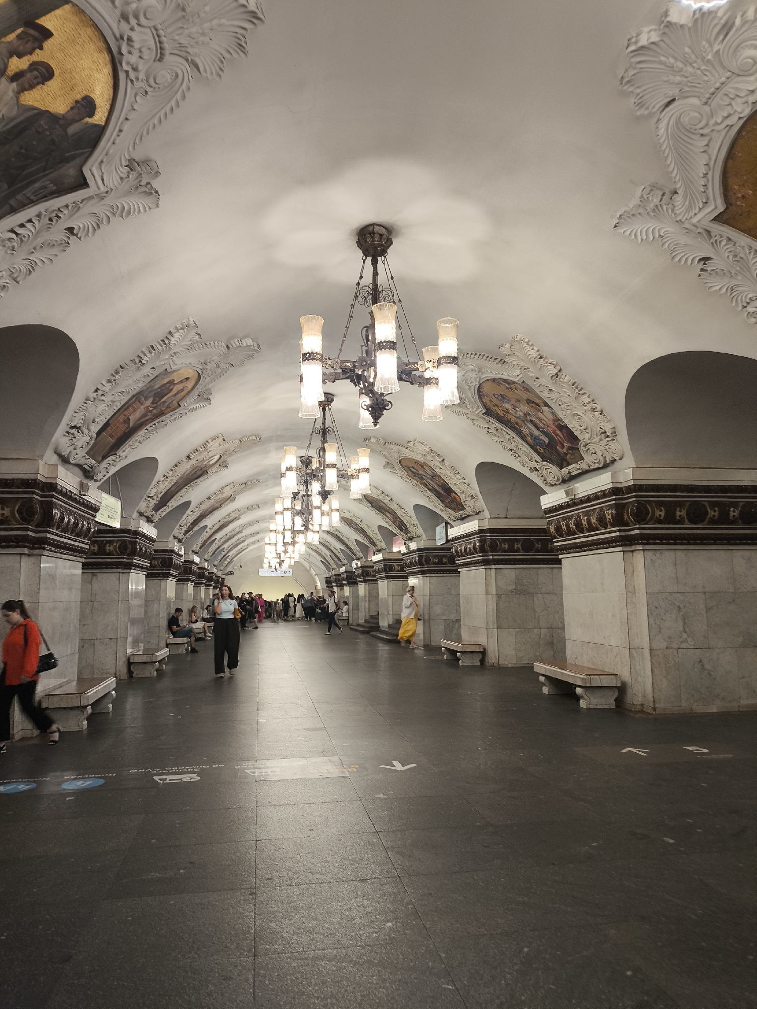 The image depicts an interior scene of a grand, ornate subway station, likely in a European city given the architecture. The station features a high, vaulted ceiling adorned with intricate white stucco work and large, elaborate chandeliers that emit a warm, bright light. Along the curved walls, above the marble-clad pillars, are framed paintings or mosaics depicting figures in historical or allegorical scenes. Marble benches are placed along the platform for waiting passengers.
Several people are visible on the platform. In the foreground on the left, a woman in an orange jacket and black pants walks away from the camera, carrying a black bag. Further down the platform, a woman in dark pants and a light blue top is walking towards the camera. Other figures are scattered further down the platform, some sitting on benches and others walking, suggesting moderate passenger activity. One woman in a yellow dress stands to the right, looking away.
The floor is dark gray and appears to be made of polished stone. Some white directional arrows are visible on the floor, indicating pathways. The lighting is artificial, casting a warm glow throughout the station and highlighting the architectural details. The overall atmosphere is one of grandeur and historical significance, with a sense of everyday public transit use. There is no indication of the time of day or weather outside.