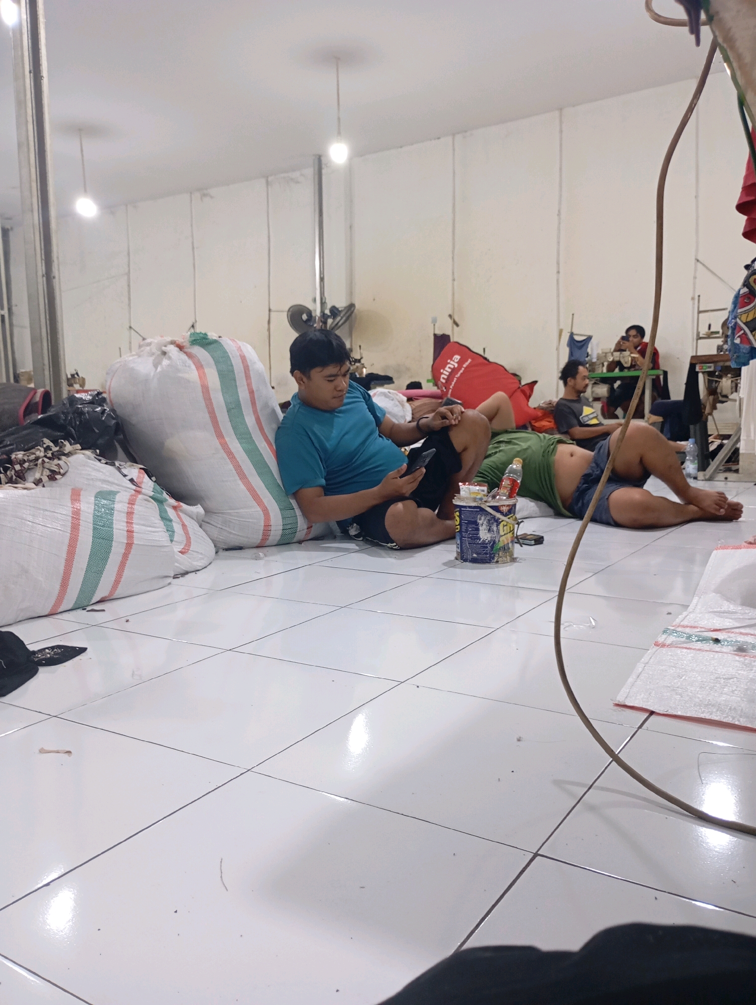 The image depicts an indoor scene, likely a workshop or storage area, with a tiled floor and white walls. Several large white sacks with green and red stripes are prominent in the foreground and midground. In the midground, a man in a blue t-shirt and black shorts is sitting on the floor, looking at his phone. To his right, another man is lying down, wearing a green t-shirt and blue shorts. A blue bucket with labels and various items is placed near the lying man. In the background, two other individuals are visible, one sitting at a table with what appears to be a sewing machine. A red object with the word "ninja" printed on it is also present in the background. The overall impression is a casual, possibly work-related environment where individuals are taking a break. The specific location is the city of Tasikmalaya, Indonesia.