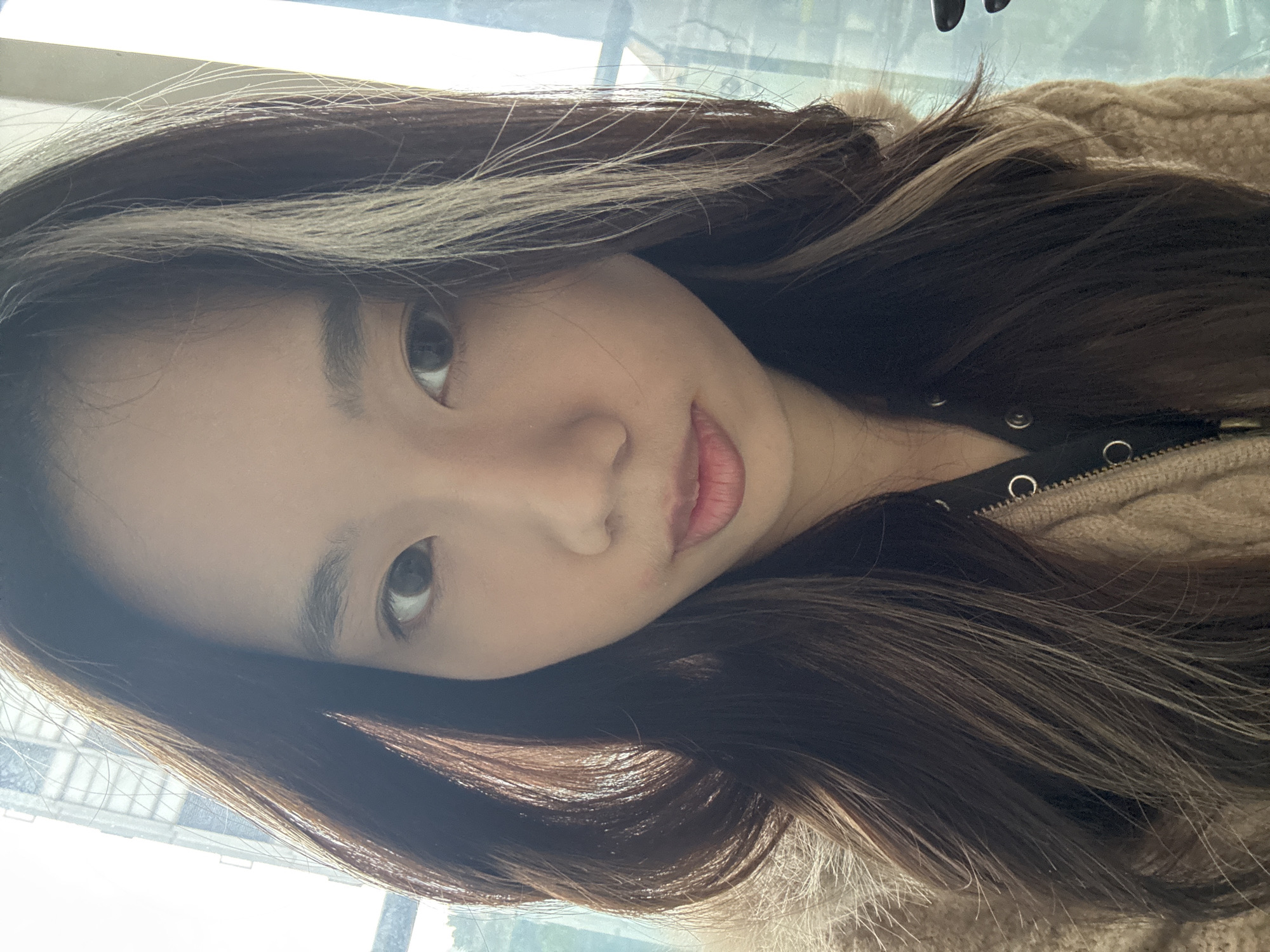 A single individual, a young adult female, is positioned centrally in a medium-close-up shot, facing directly towards the camera. She has dark eyes, dark eyebrows, and a fair complexion. Her lips are closed with a reddish tint. Her long, dark brown hair, featuring lighter highlights, frames her face and extends over her shoulders. She wears a textured light brown or beige outer garment with a zipper, over a dark underlayer that exhibits small metallic eyelets or buttons near the neckline. The background consists of an indoor setting near a window, with bright natural light. A blurred high-rise building with discernible window structures is visible through the window on the left, against a bright sky, suggesting an urban environment. A window frame or vertical support is partially visible on the right. The specific location within Nan'an District, China, is not visually identifiable from the image content. The individual is static, looking directly at the camera.