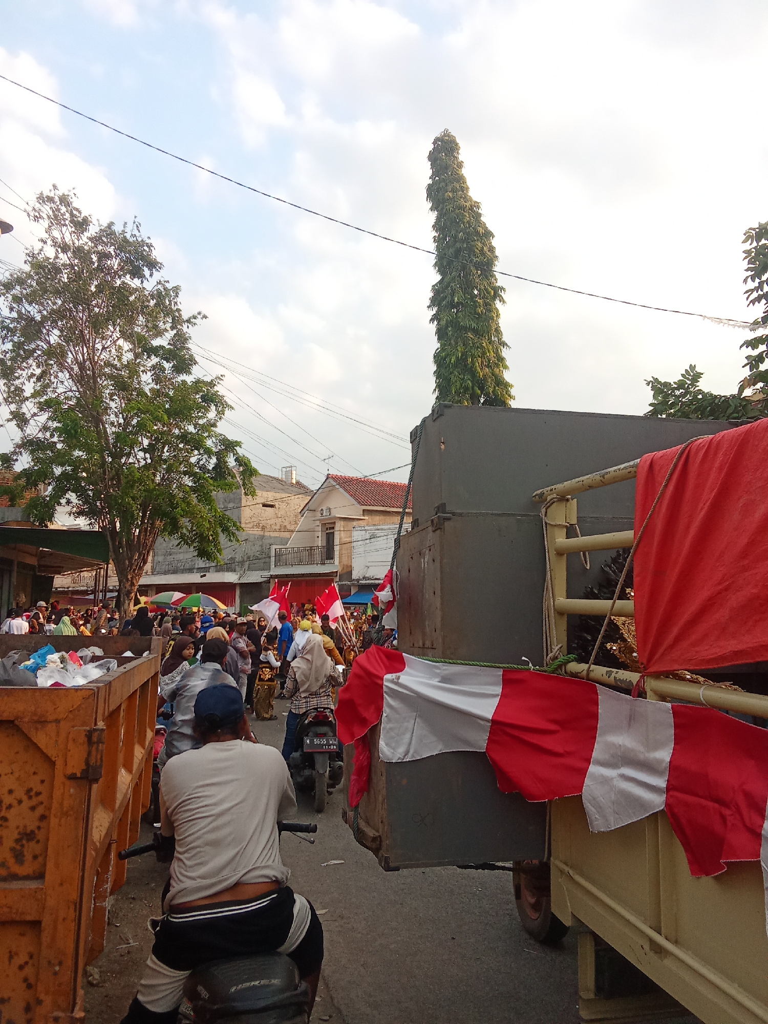 In this outdoor scene, a group of people are gathered on a street. In the foreground, a man is riding a motorcycle, facing away from the camera. He is wearing a light-colored t-shirt and black and white shorts. To his left, a large, rusty orange dumpster is filled with trash. To his right, a truck is visible, adorned with red and white fabric banners. The truck appears to be carrying a large, dark gray box.
In the middle ground and background, a crowd of people is moving along the street, many holding red and white flags. The street is lined with buildings, some with red roofs. A tall, slender tree with dense green foliage stands to the left of the frame. The sky above is partly cloudy. The overall impression is of a public gathering or procession.