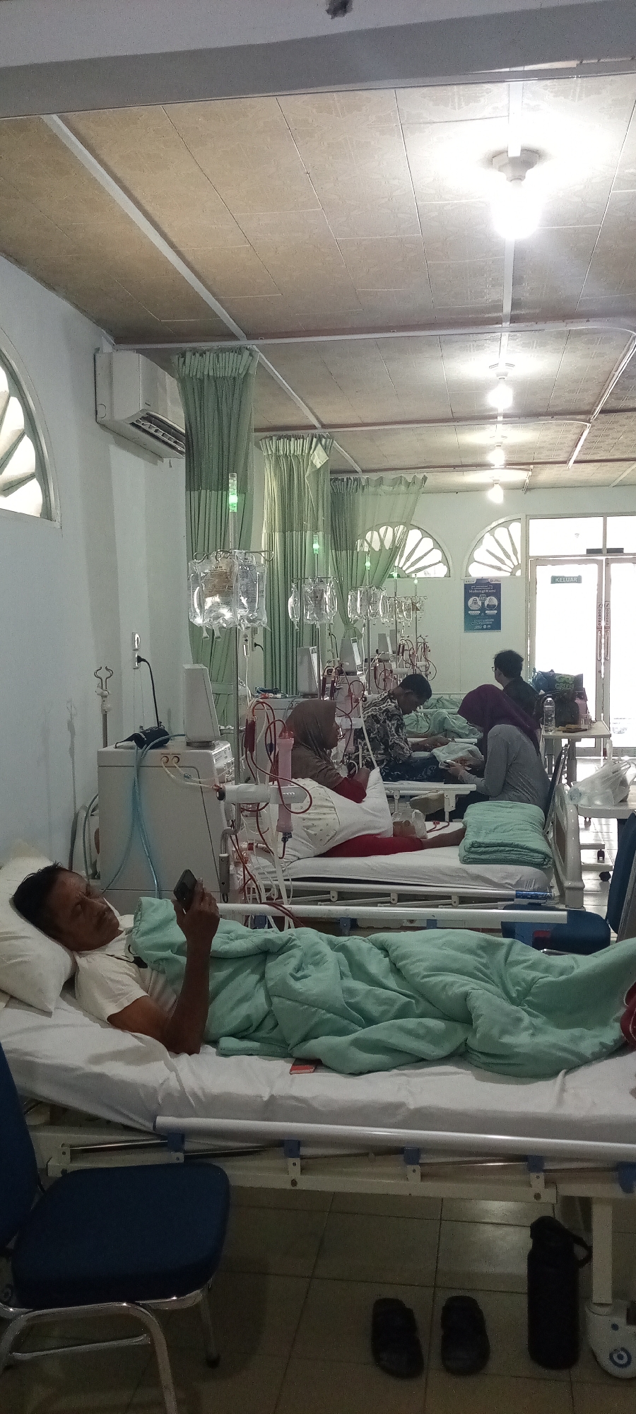 The image depicts an interior scene, likely a hospital ward, in Lhokseumawe, Indonesia. The room is lit by overhead fluorescent lights and natural light from arched windows with decorative grilles. The ceiling is a textured, beige tiled suspended ceiling with visible metal framing and light fixtures. Green curtains hang along the walls, some partially obscuring windows.
In the foreground, a man lies on a hospital bed, covered by a light green blanket. He is propped up on pillows and appears to be holding a mobile phone in his right hand, looking down at it. He is wearing a white t-shirt. The bed is a standard hospital bed with a metal frame.
Behind him, several other hospital beds are visible, each occupied by a patient. These patients are also covered by blankets and appear to be undergoing medical treatment, as indicated by the presence of dialysis machines and IV drips. Several individuals, possibly medical staff or other patients, are tending to or sitting with the patients. One individual in the middle ground, wearing a patterned shirt, is interacting with a patient. Another individual wearing a hijab is seated next to a patient.
Medical equipment, including dialysis machines with tubing and bags of fluid, is prominently displayed throughout the ward. The floor is tiled with large, light brown tiles. On the floor near the foreground bed, a dark blue chair and a pair of black flip-flops are visible. Further back, a black water bottle and another pair of flip-flops are also present. A sign with text in Indonesian, including the word "KELUAR" (exit), is visible on a door in the background. The overall atmosphere suggests a clinical and somewhat sterile environment, with an emphasis on patient care.