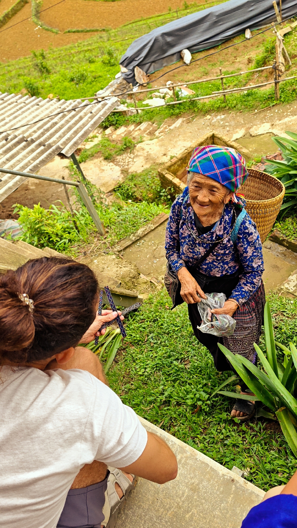 This overhead shot depicts an interaction between two individuals in a lush, rural setting identified as Dế Xu Phình, Vietnam. In the foreground, viewed from behind, a person with dark hair tied in a bun with a silver clip sits or crouches on a concrete step. This individual wears a light grey short-sleeved t-shirt, grey shorts, and light-colored Keen brand sandals. Their right hand extends forward, holding a patterned, dark-colored beaded item, possibly a bracelet or decorative craft.
Facing this individual, slightly to the right, is an elderly woman, standing on a dirt and grass path. She is smiling broadly, revealing deep facial wrinkles, and wears a vibrant plaid headwrap composed of blue, green, pink, and red squares. Her attire consists of a long-sleeved blue top with a floral pattern, a dark skirt featuring an intricate geometric pattern along the hem, and dark sandals. A large woven basket is visible on her back, and a small dark bag is slung over her left shoulder. Her hands are clasped around a clear plastic bag containing multiple small, colorful items.
The immediate environment around them consists of varied green grasses and larger leafy plants. A concrete ledge runs across the mid-ground. To the upper left, a grey corrugated metal roof of a structure is visible. In the background, rolling hills with distinct brown terraced agricultural fields, possibly rice paddies, are prominent. A dark grey tarp covers a mound of earth or debris further back, secured by wooden posts. The overall scene suggests a cultural exchange, possibly a transaction, amidst a verdant, mountainous landscape.