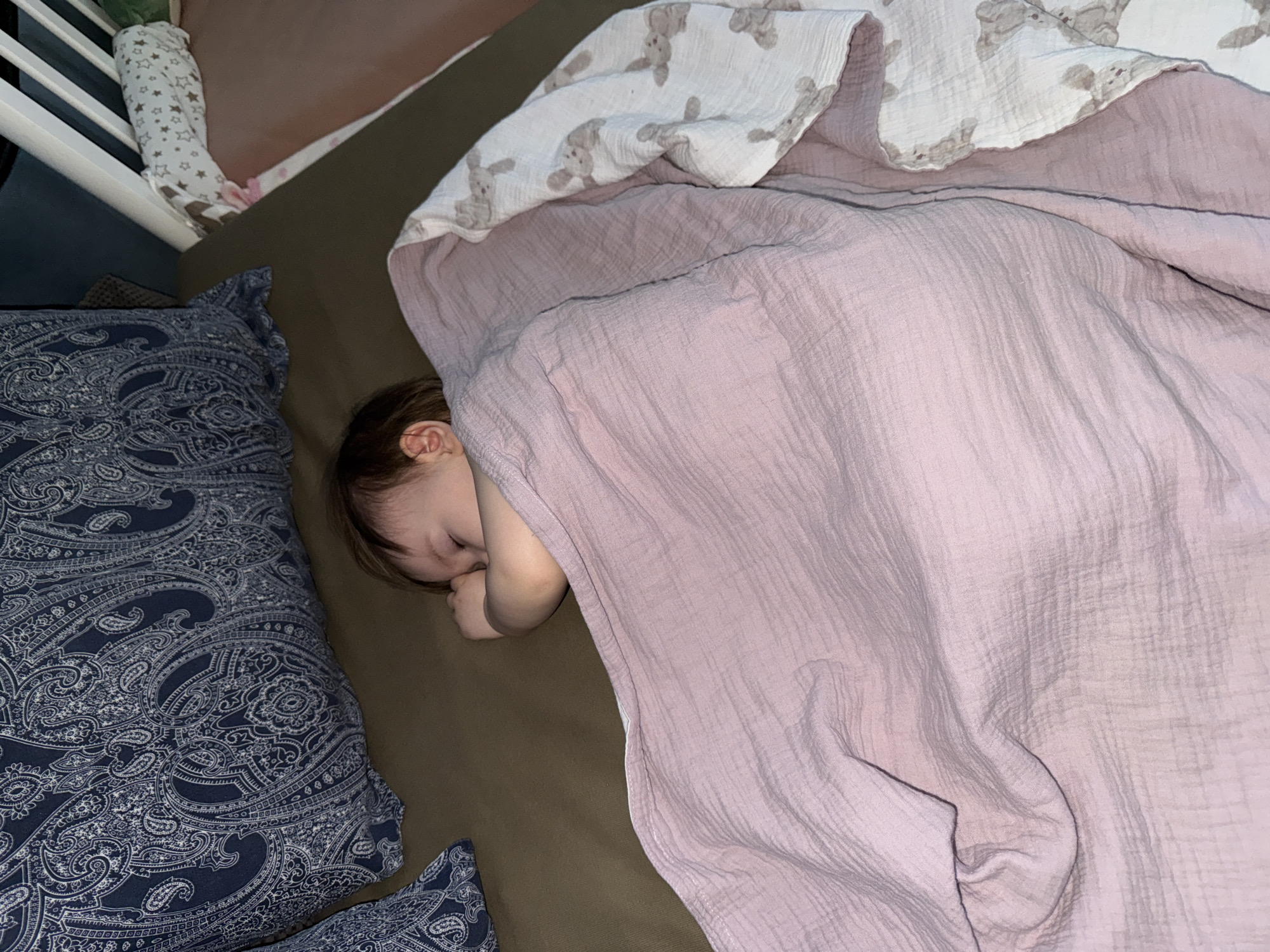 A child sleeps in a bed. The child is covered by two blankets: a light pink blanket and a white blanket with a bunny pattern. A blue paisley pillow is positioned next to the child's head. Part of a white crib is visible on the left side of the image. The location is not evident from the image.