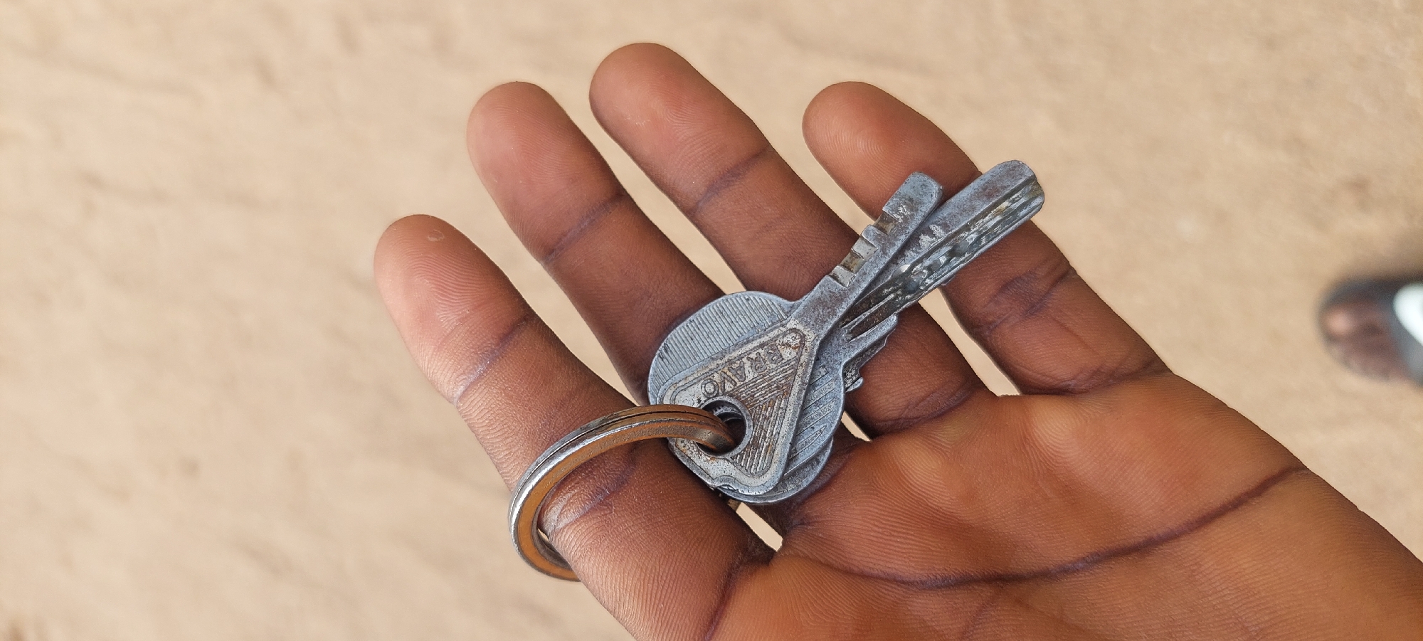 A close-up, top-down shot of a person's hand holding two metal keys on a silver keyring. The hand appears to be dark-skinned with visible wrinkles and creases, specifically on the fingertips and palm. The keys are aged and slightly worn, with one key appearing to be slightly longer and more detailed than the other. The keyring is a simple, circular silver band. The background is a textured, light brown surface, possibly sand or dry earth. The image is taken in Jalingo, Nigeria. A blurry image of a shoe can be seen at the bottom of the frame.