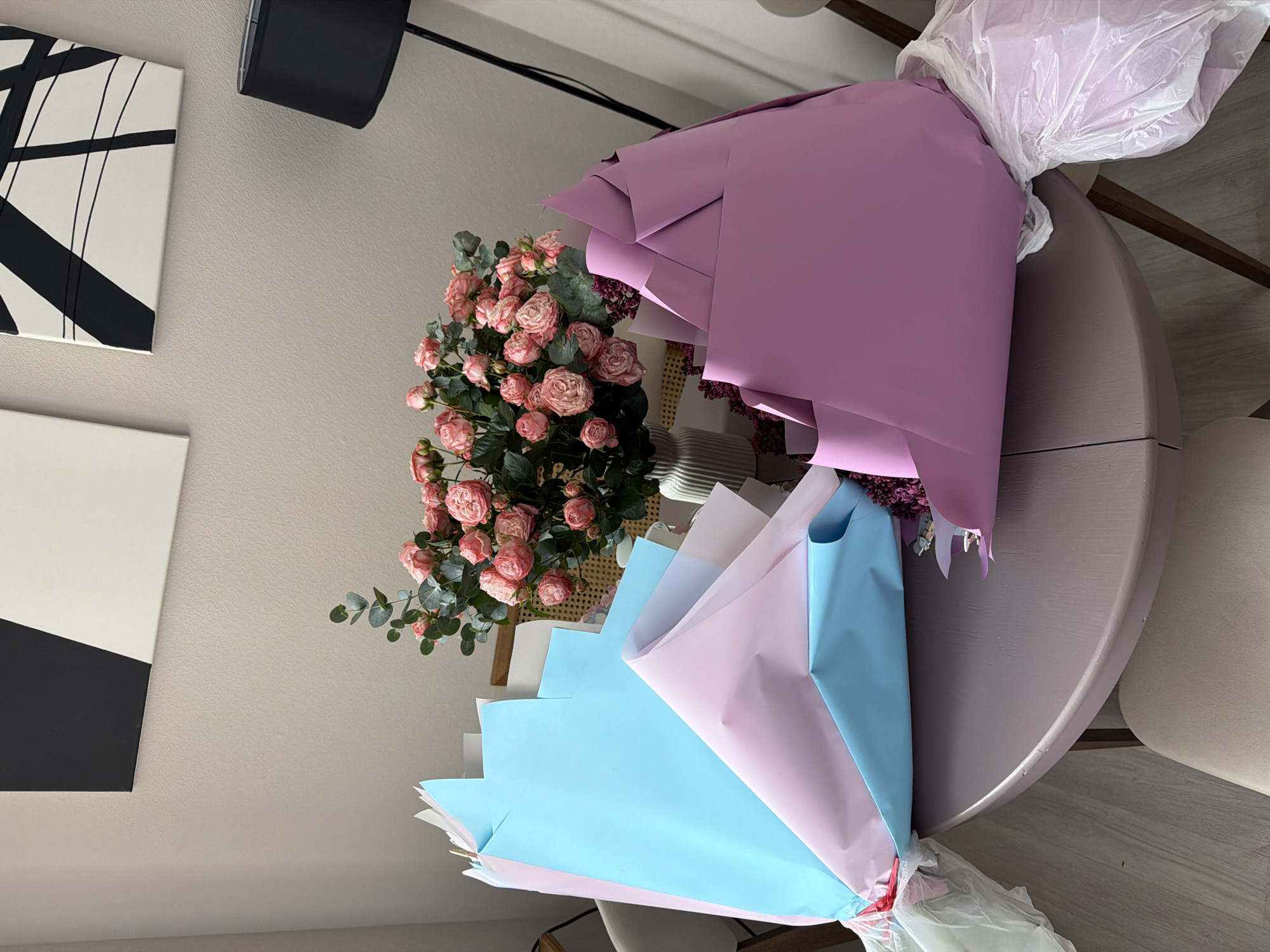 On a round, lavender-colored table, several bouquets of flowers are arranged. One bouquet features pink roses with green foliage. Two additional bouquets are wrapped in colored paper: one in lilac and the other in alternating layers of light blue, white, and pink paper. All bouquets appear to be tied at the base with plastic and ribbons. The table sits on a wooden floor. Adjacent to the table, there is a black speaker, and hanging on the wall are two pieces of art that are black and white. The environment is indoors with neutral-toned walls. It is unclear if this location is in Moscow, Russia.