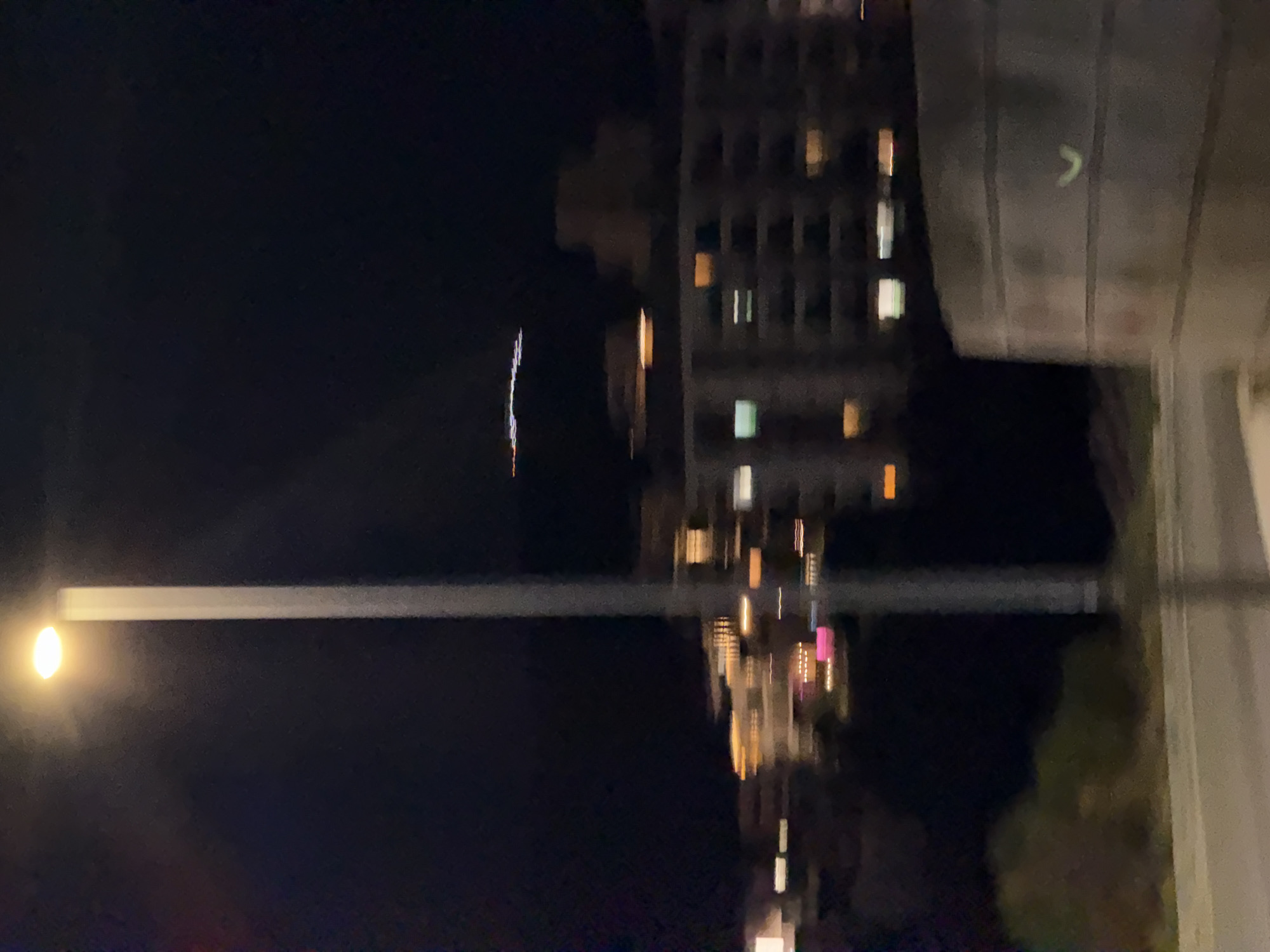 The image depicts a nighttime scene in Boreti, Montenegro. A tall, grey lamppost with a bright, illuminated light is positioned prominently in the foreground, casting a warm glow. The background is a blurry panorama of city lights, suggesting buildings and structures. The overall impression is one of a moderately populated urban area at night, with artificial light sources dominating the scene.