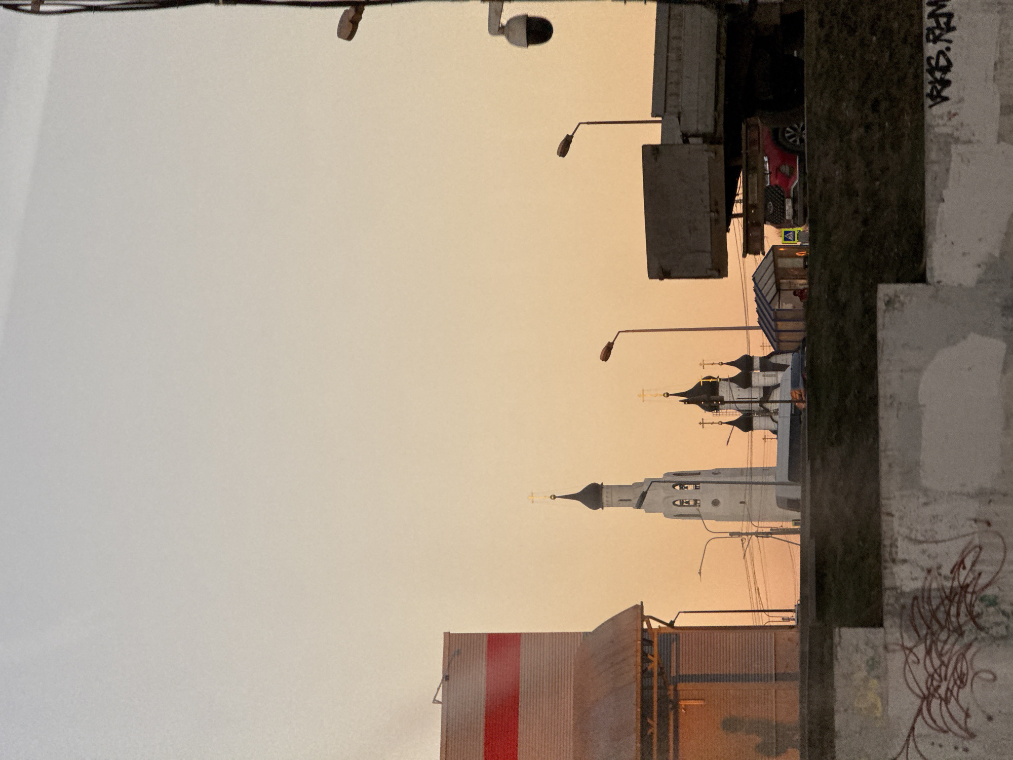 The image shows a street view in Saint Petersburg, Russia, at dusk or dawn, characterized by a predominantly orange sky. In the foreground, there is a concrete wall with graffiti. Buildings, including one with a red and white vertical stripe, and a church with multiple towers topped with crosses, are visible in the middle ground. Street lamps line the scene, and overhead wires are strung across the sky. In the upper part of the image, there is a truck, and a traffic sign is seen near the building.