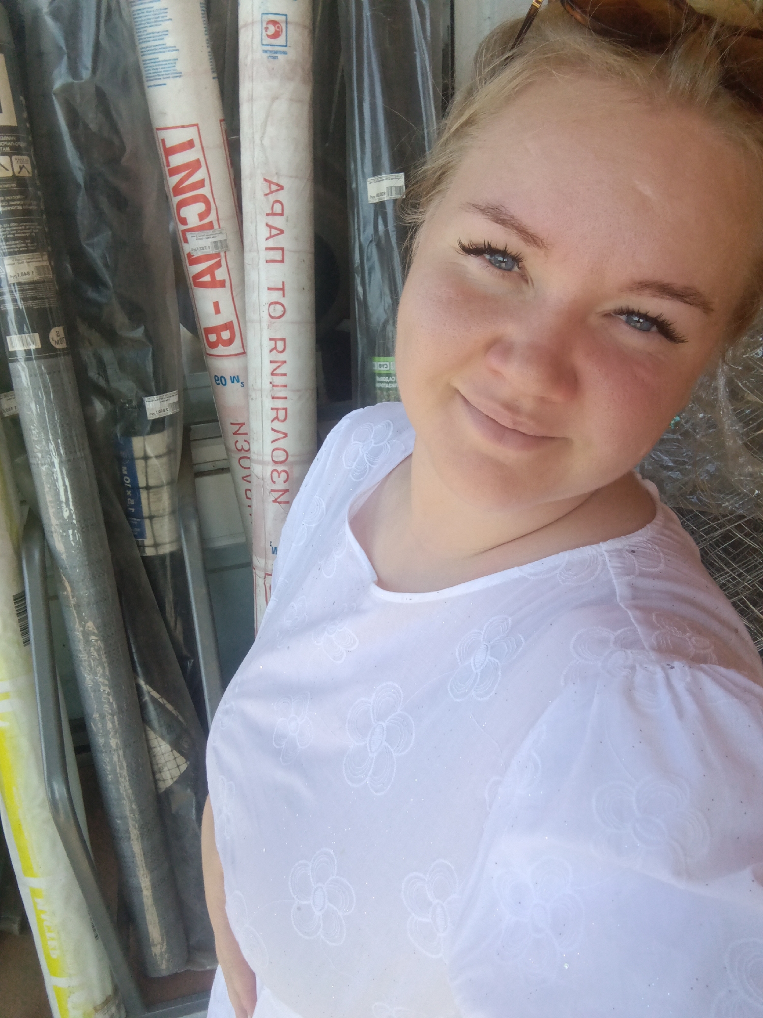 A blonde woman with blue eyes smiles into the camera, her head tilted slightly to the left. She is wearing a white top with embroidered flowers and sparkly accents. Behind her, to the left, are several large rolls of material, some wrapped in plastic and others with visible labels. One label reads "INCLIV-B". To the right of the woman, more rolls of material are visible, and the scene appears to be outdoors, possibly in a storage area or market in the city of Fokino, Russia.
