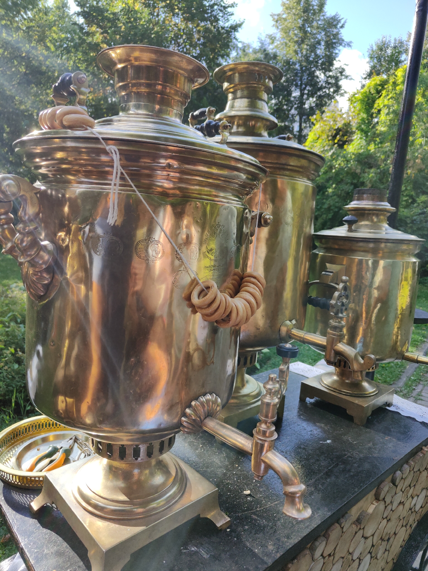 The image depicts an outdoor scene with three brass samovars of varying sizes arranged on a dark stone surface. The samovars are ornate and polished, reflecting the sunlight. A string of dried bagels, known as bubliks, is draped over the largest samovar. In the foreground, a gold-colored tray with what appear to be pliers and a green object rests on the stone surface. The background features lush green trees and foliage, suggesting a park or garden setting. The sky is visible, with some clouds. The overall impression is a still life arrangement of traditional Russian tea-making equipment in a natural environment. The specific location within Moscow, Russia, cannot be determined from the image alone. There are no people or animals visible in the scene.