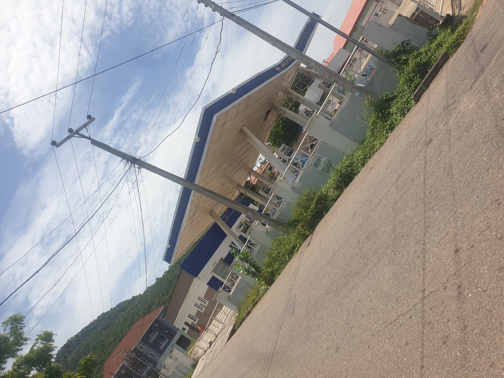 The image shows a street scene in Lokoja, Nigeria. On the left, there are utility poles with electrical wires running across the sky. In the background, a green, tree-covered hill is visible. A "JOELATOS" branded filling station is located on the side of the road. A low concrete curb runs along the edge of the pavement, with some vegetation growing along it. Another building is further back from the filling station and appears to be residential. The pavement is light brown.