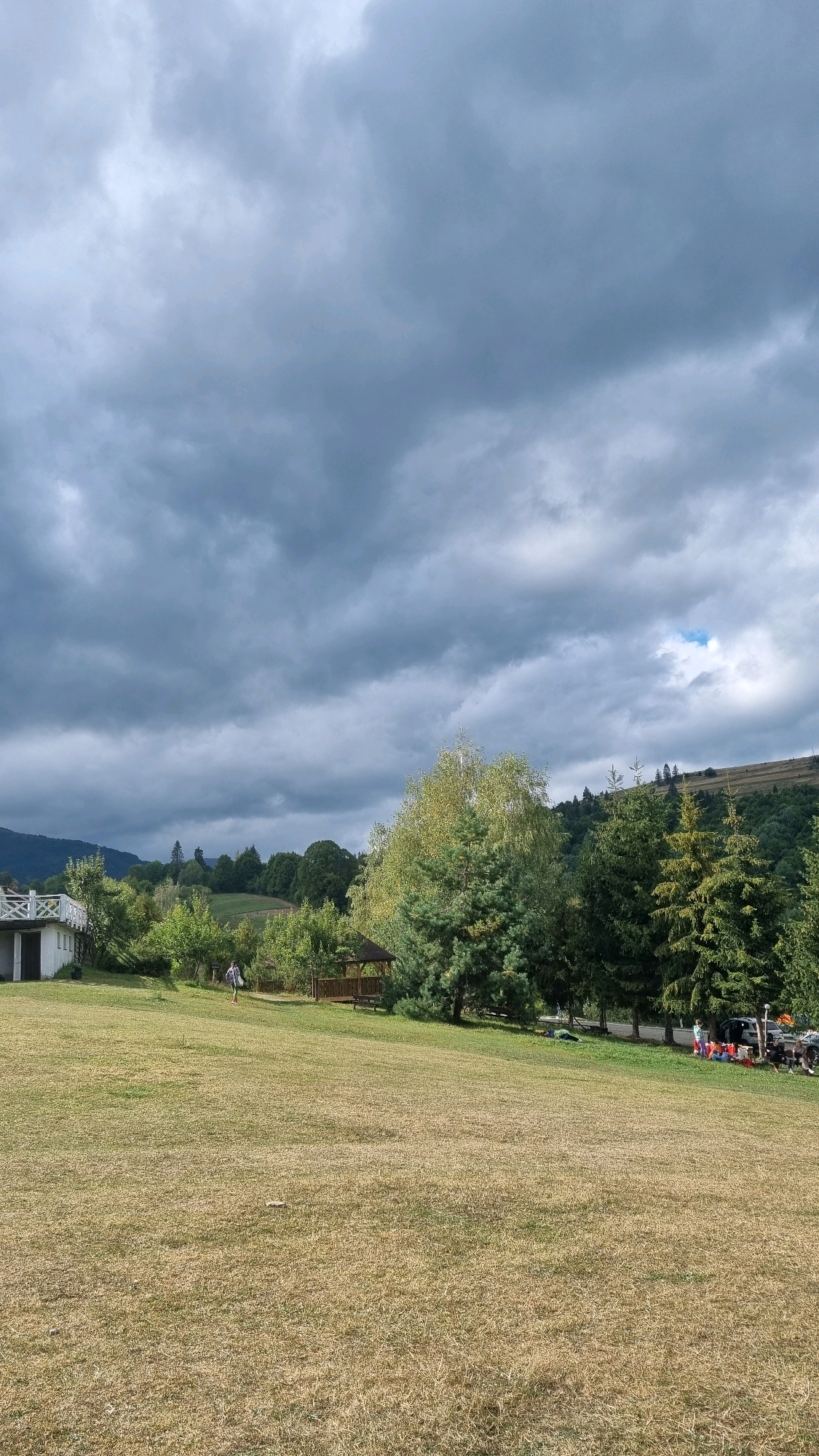 This image depicts a grassy hillside in Izky, Ukraine, under a cloudy sky. In the foreground, a dry, mowed lawn stretches towards a line of trees and a building. To the left, a white house with a porch is visible. In the middle ground, a wooden gazebo sits on the lawn, and a person in athletic attire is walking away from it. Further into the background, a dense forest covers the rolling hills, with some trees displaying autumnal colors. A few cars and people are gathered on the right side of the image, suggesting a park or recreational area. The overall atmosphere is serene, with the overcast sky creating a subdued mood.