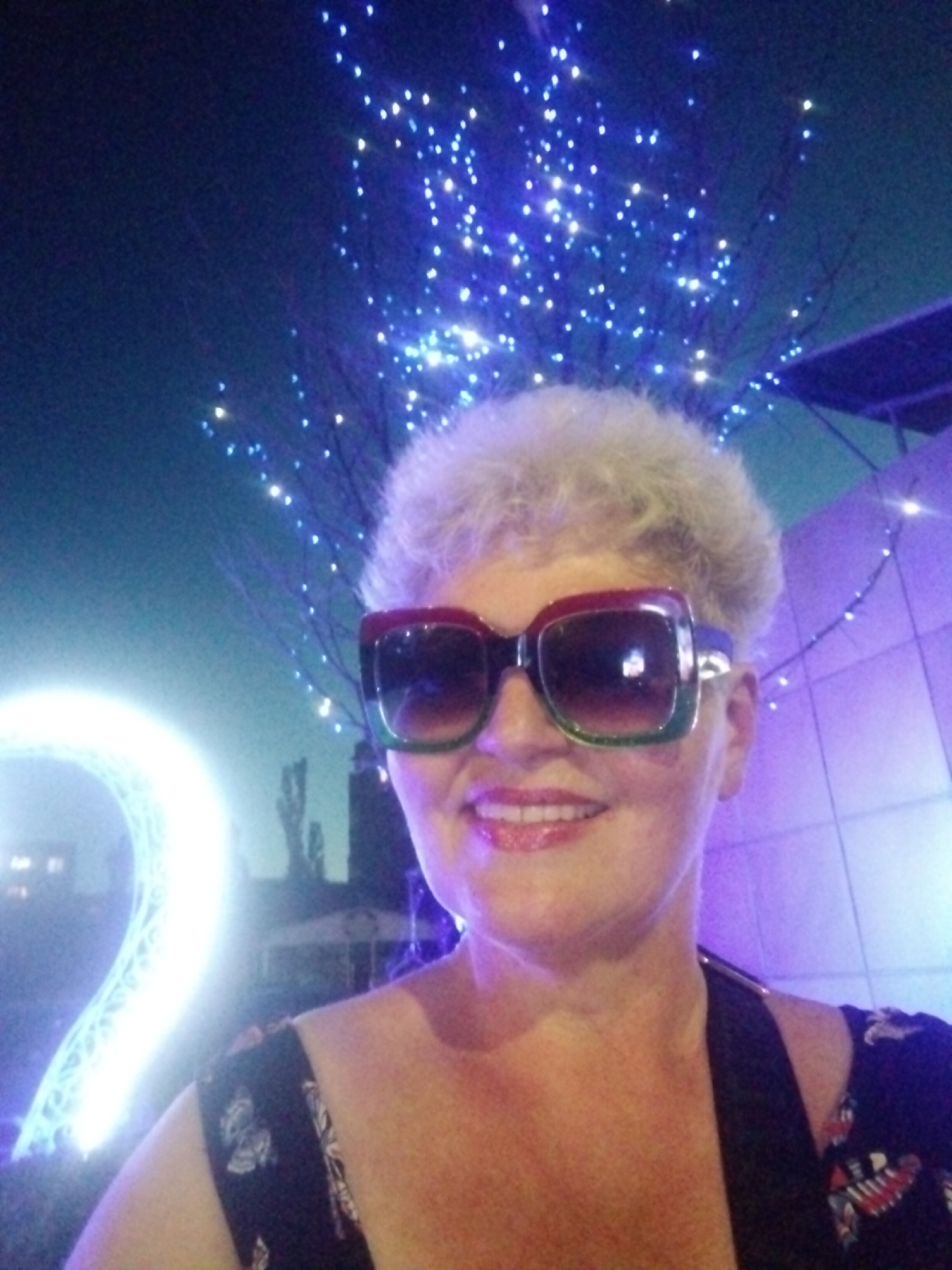 A woman with short, blonde hair and large, colorful-framed sunglasses smiles at the camera. She is wearing a dark-colored top with a floral pattern. Behind her, a large, illuminated, spiral-shaped object glows brightly, possibly a light installation. The background is dark and appears to be outdoors at night, with numerous small, blue lights twinkling like stars or a light display. A purple-lit building or structure is visible to the right. The scene suggests an outdoor event or attraction, possibly in the city of Horishni Plavni, Ukraine, given the context of other images.