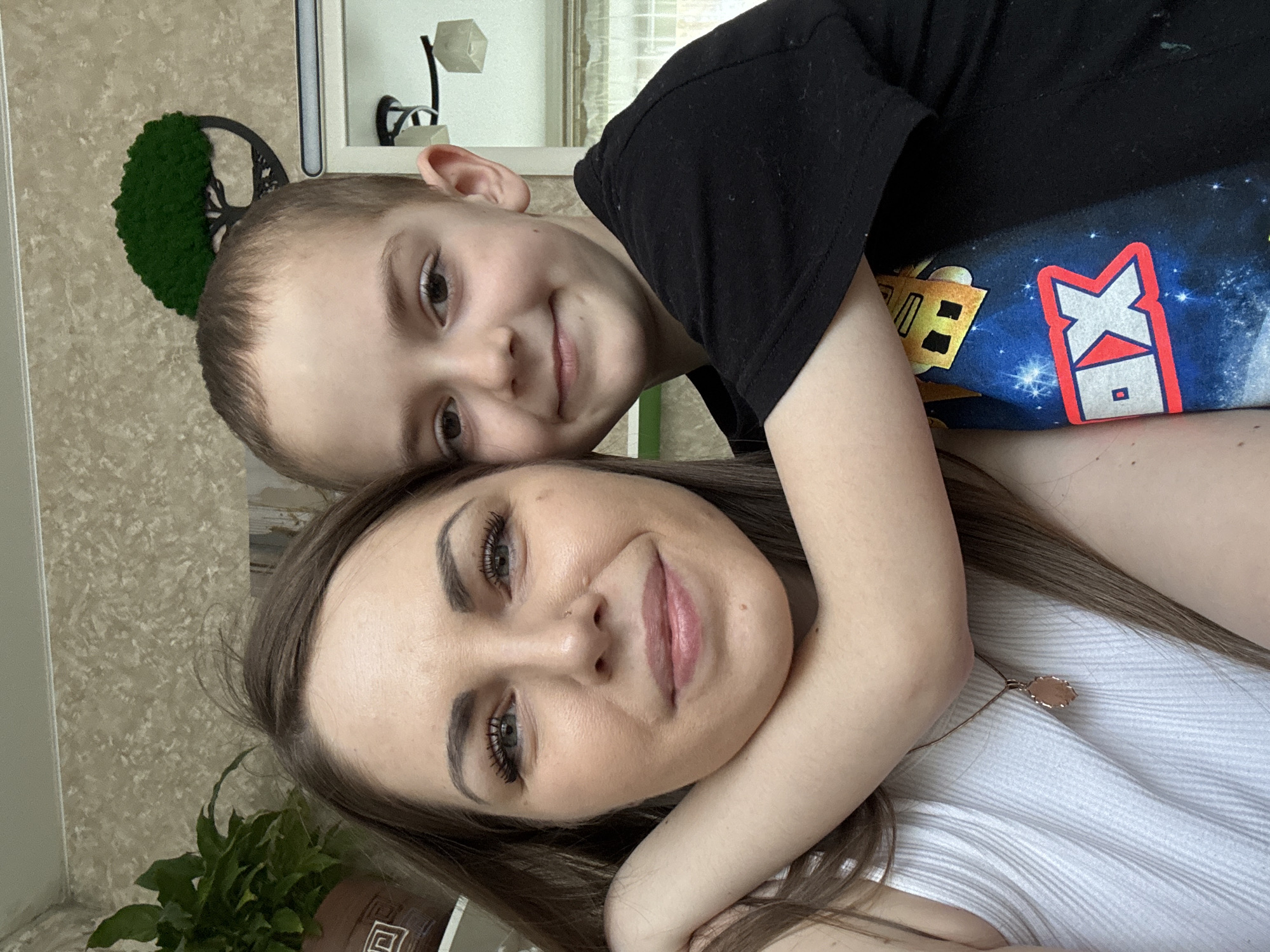 A selfie shows a woman and a child posing closely. The woman has long brown hair and wears a white ribbed tank top and a necklace. The child is leaning on her, wearing a black t-shirt with a colorful cartoon graphic. The background includes a wall with a decorative tree-shaped object, a plant, and a lamp fixture. The location is Раздолье, Russia.